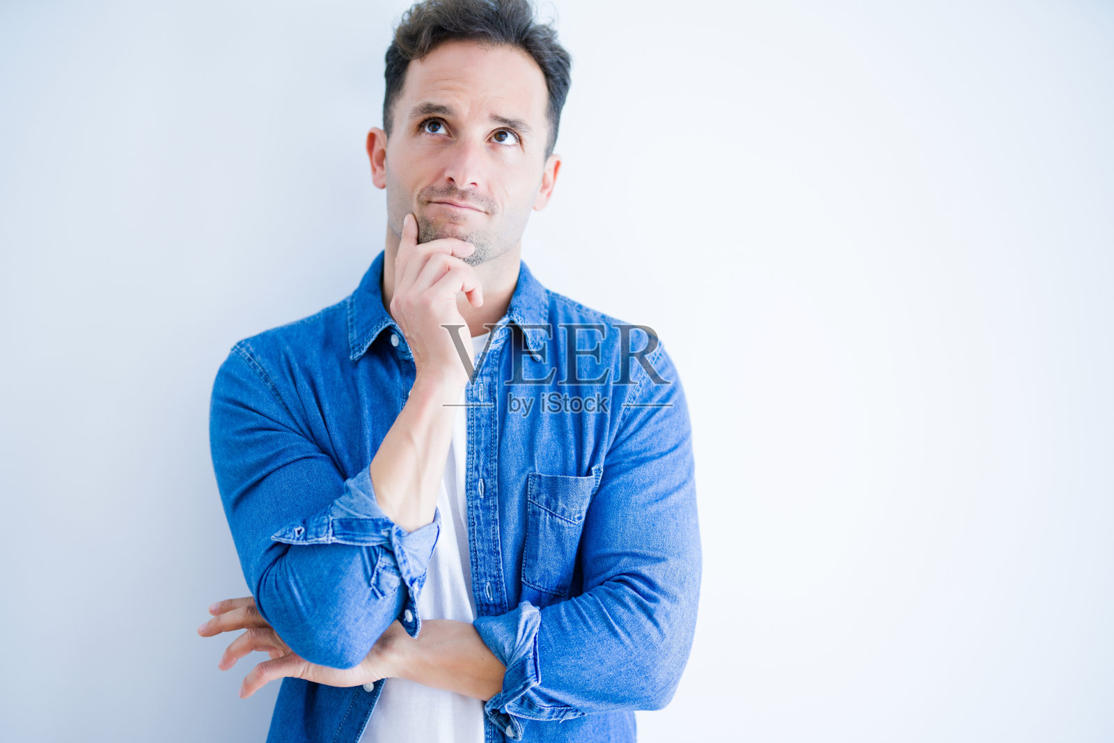 年轻英俊的男子穿着牛仔衬衫站在孤立的白色背景与手下巴思考问题,沉思的表情。带着沉思的微笑。怀疑的概念。照片摄影图片