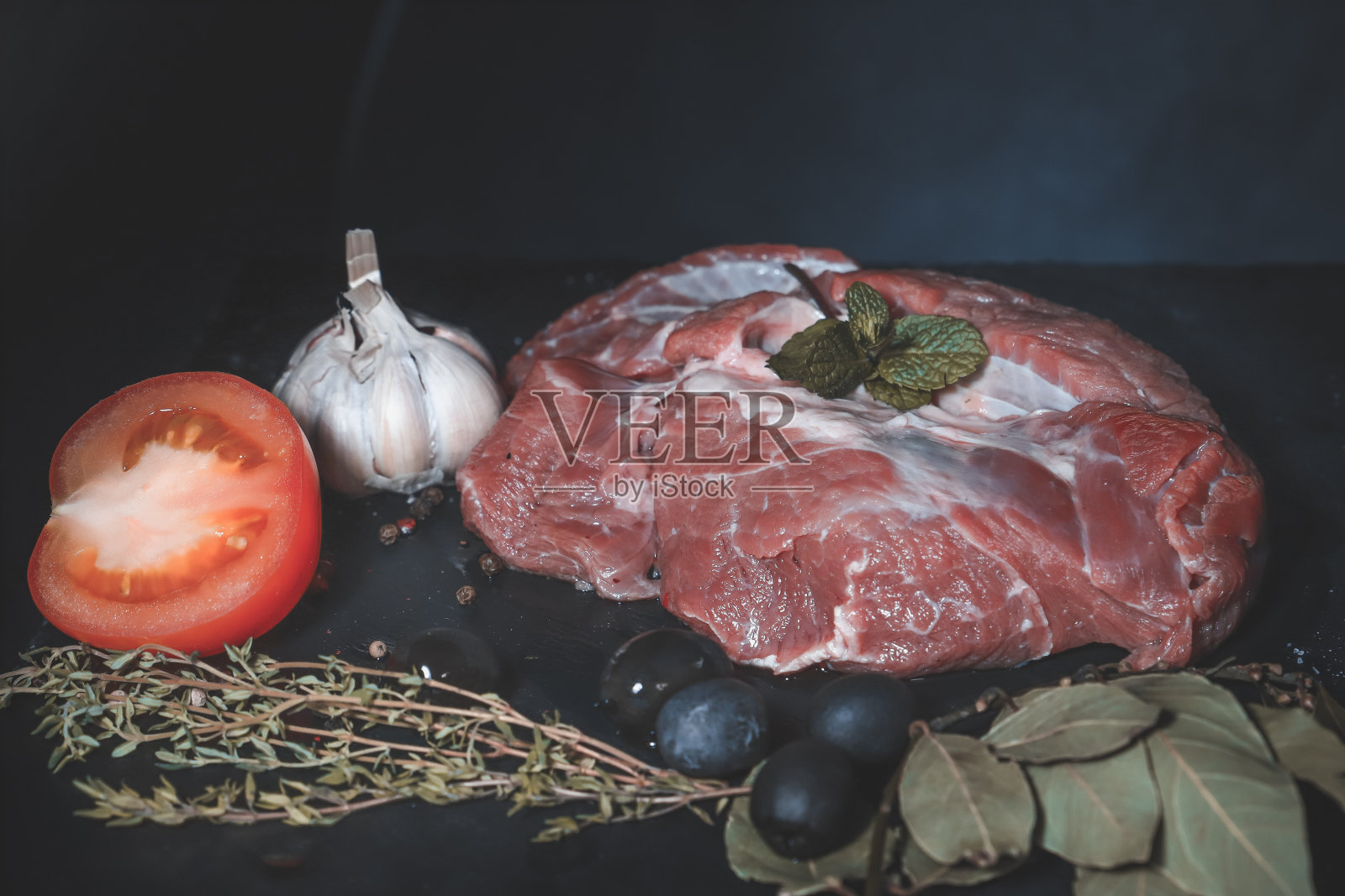 黑暗的工作台面上放着一块生牛肉,上面点缀着百里香和栗子叶,还有黑橄榄和一颗蒜头照片摄影图片