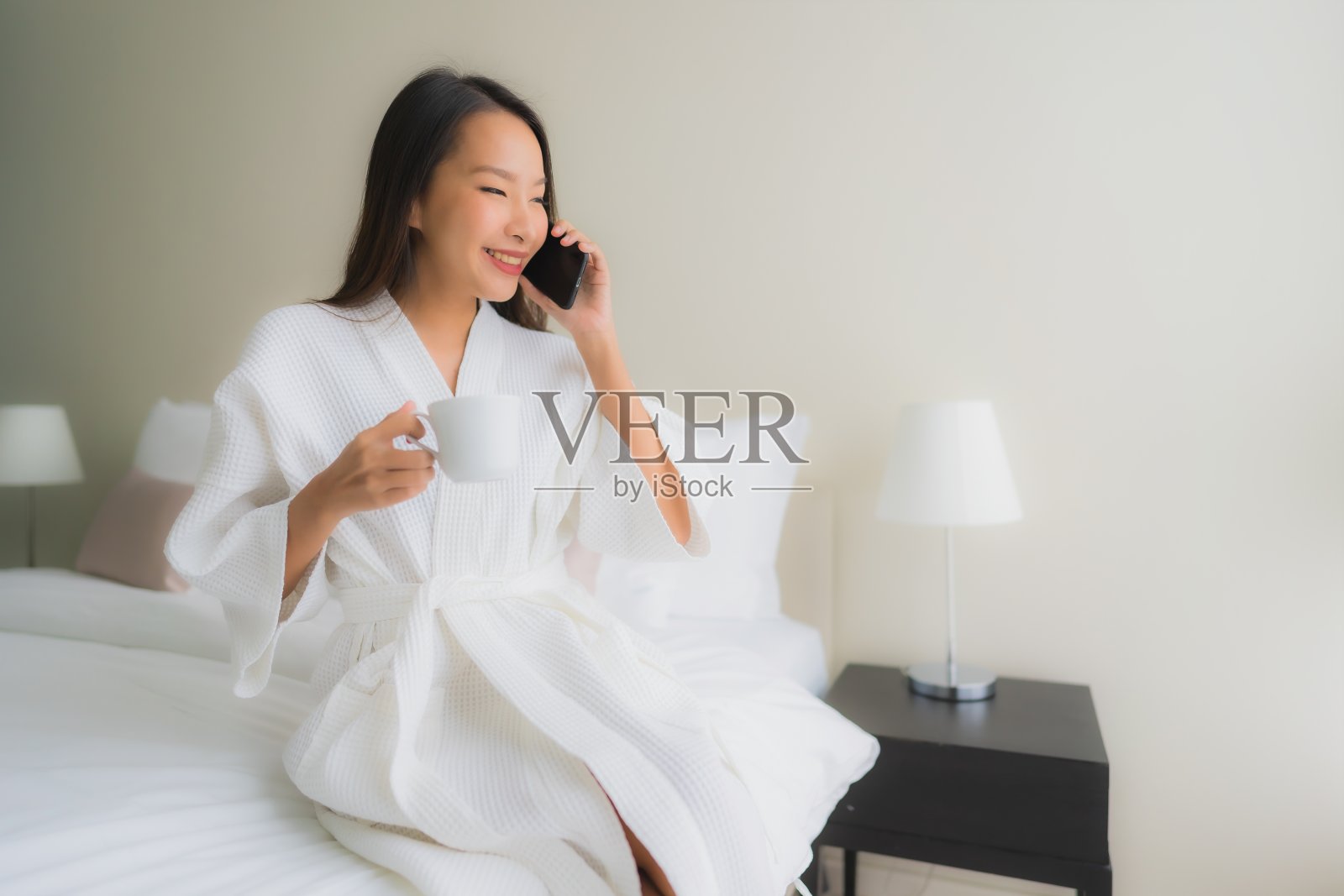 描绘美丽的年轻亚洲女性在床上拿着咖啡杯和手机照片摄影图片