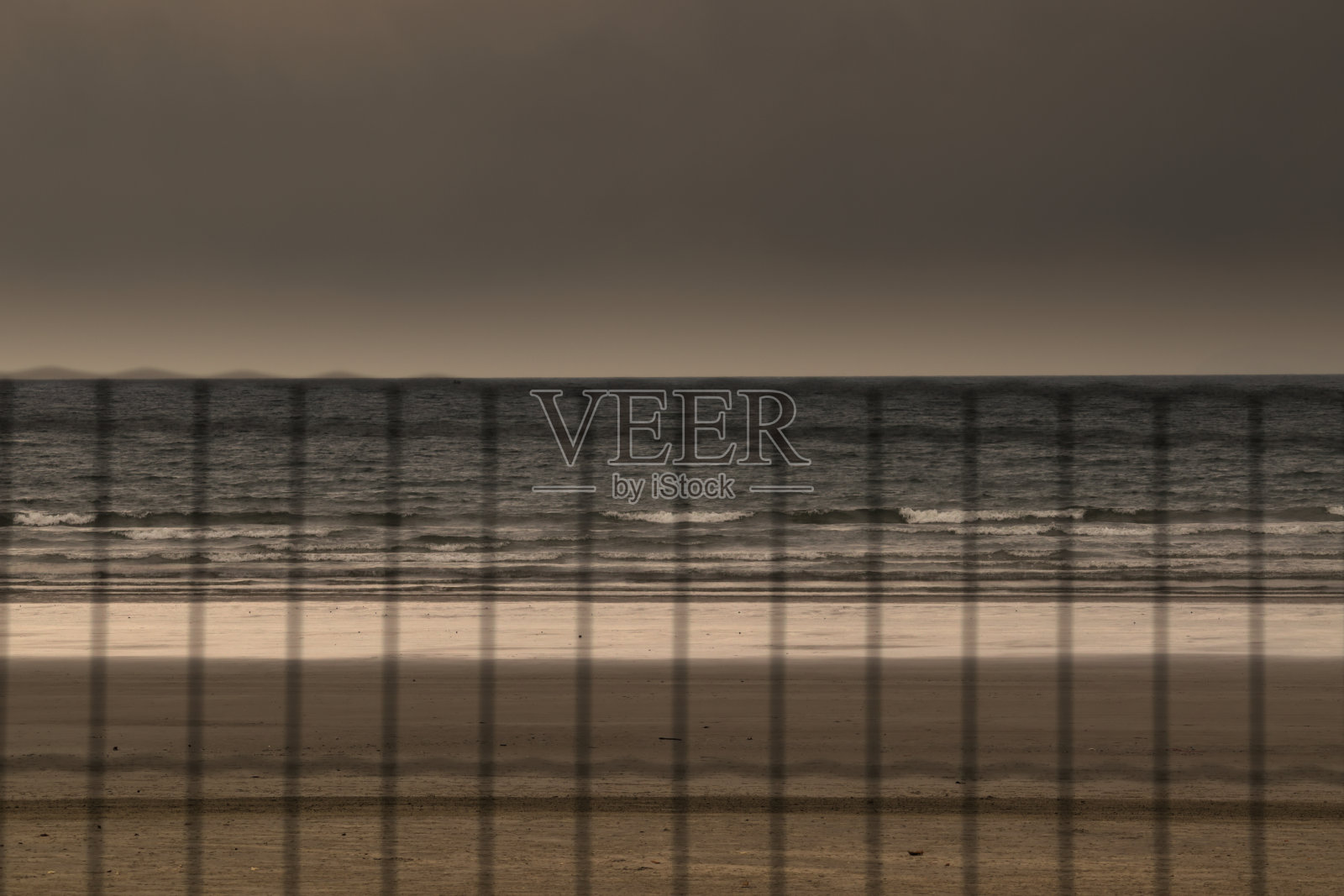 深色的沙带和灰色的海在背景中,反射着多云的天空的光,观察钢丝网的编织。照片摄影图片