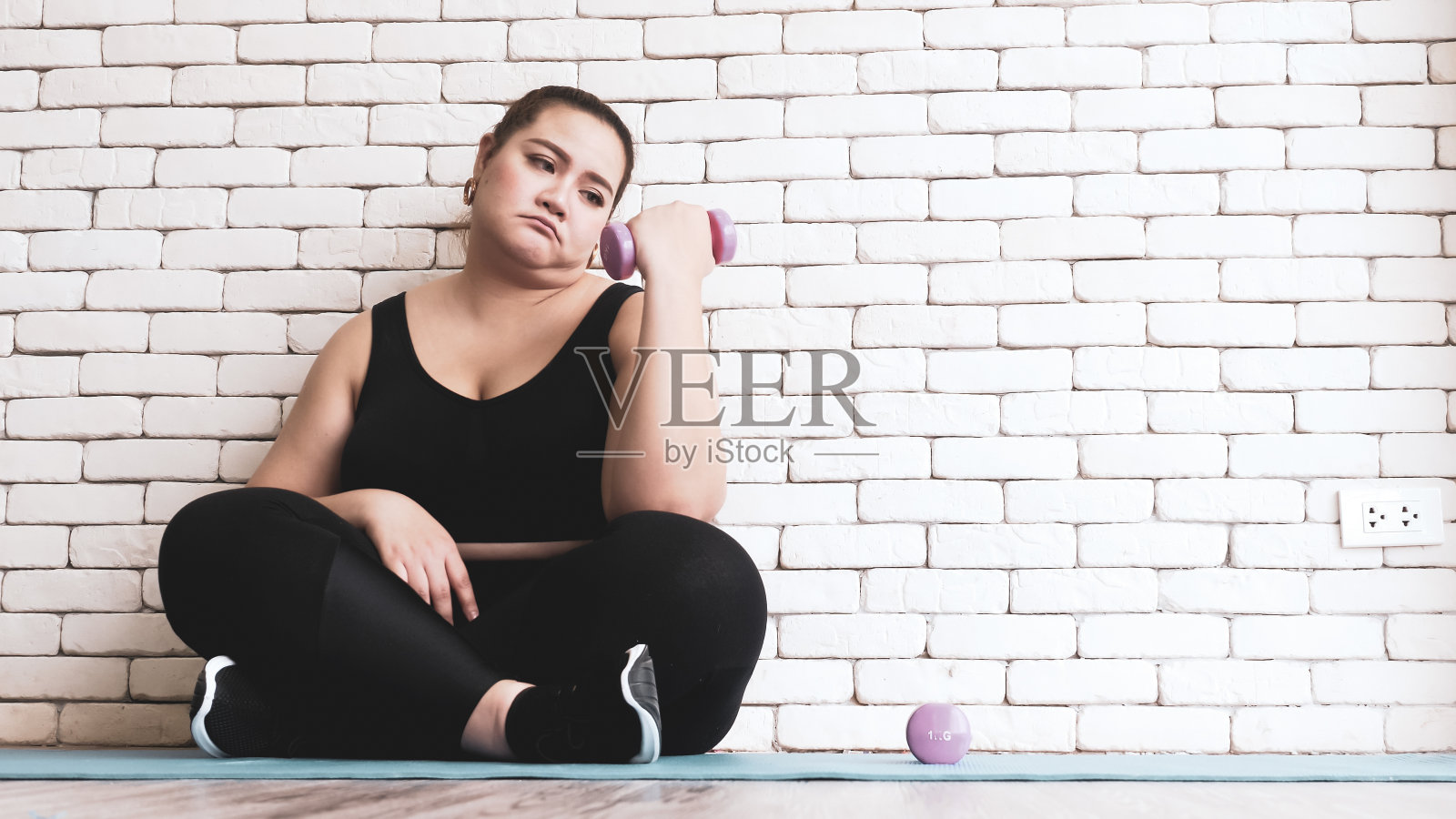 亚洲胖乎乎的女人坐在瑜伽垫上,举起哑铃感觉不开心。照片摄影图片