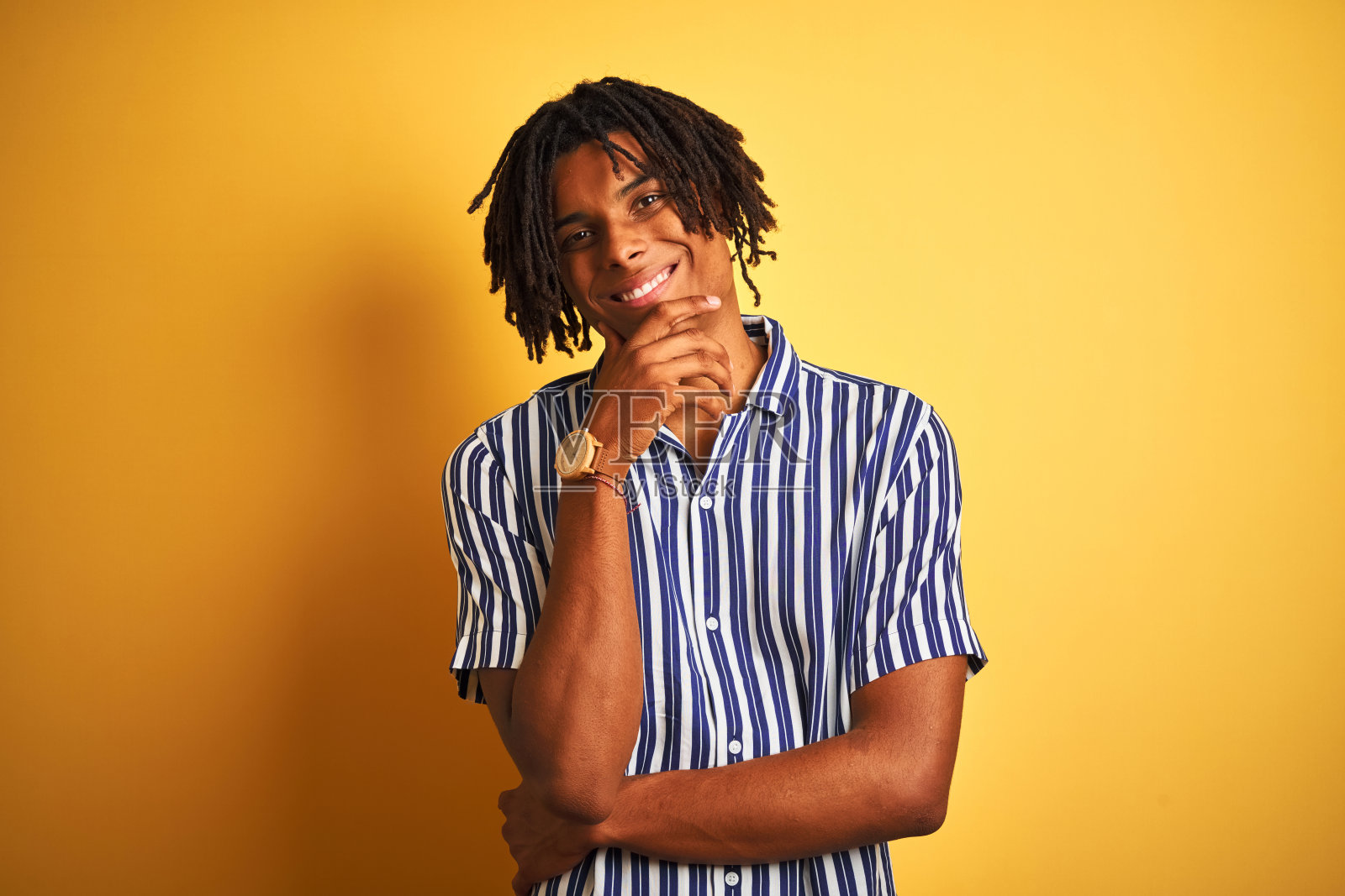 一个留着长发绺、穿着休闲条纹t恤的黑人,在孤立的黄色背景下,看起来很自信地看着镜头,双臂交叉,手举在下巴上。积极的思考。照片摄影图片