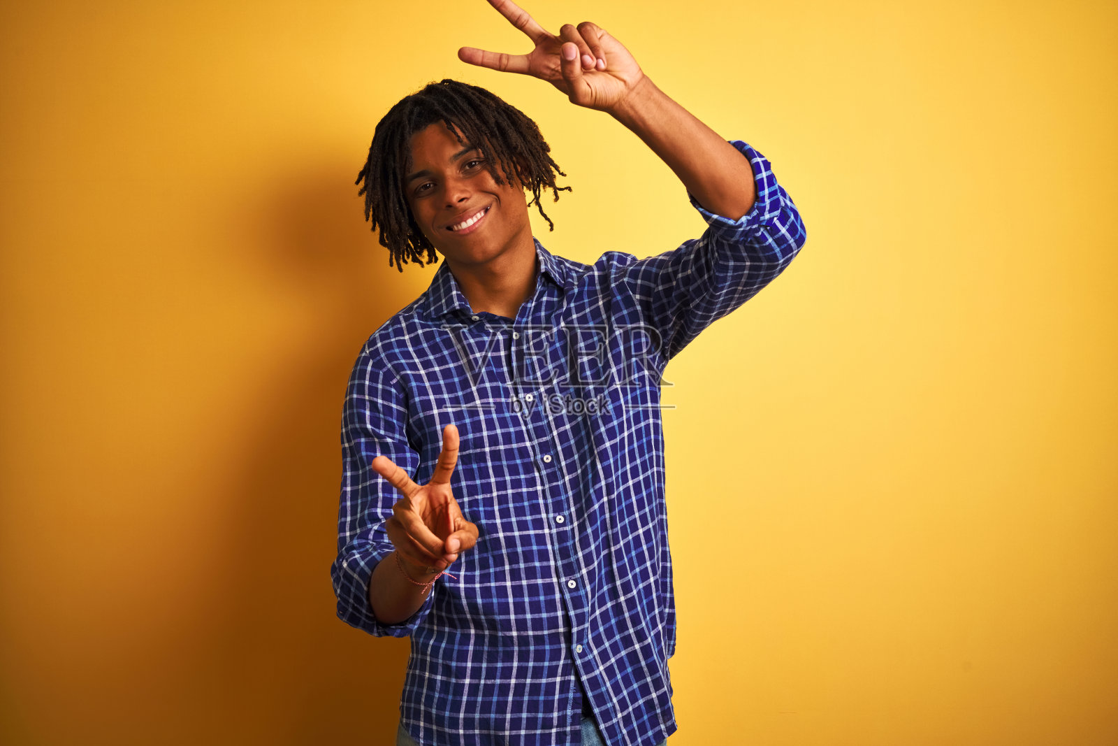 一个留着雷绺、穿着休闲衬衫的黑人站在孤立的黄色背景前微笑着看着镜头,手指做着胜利手势。二号人物。照片摄影图片