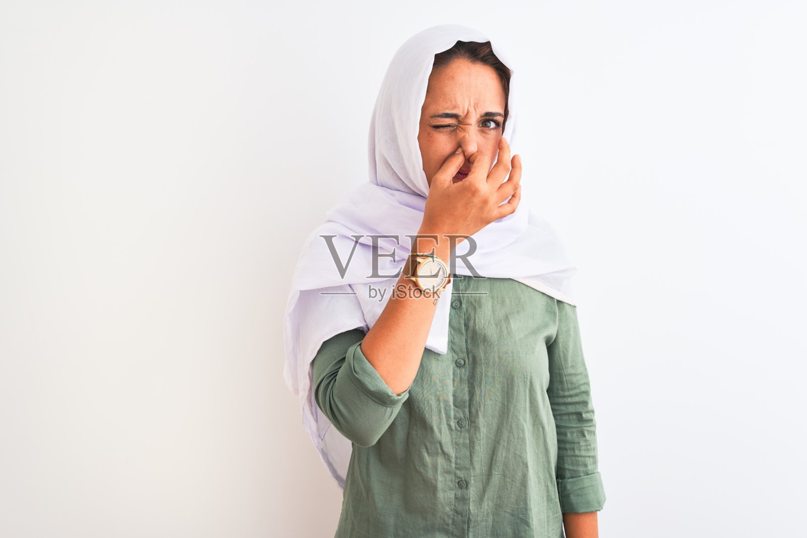 年轻美丽的阿拉伯妇女戴着传统的穆斯林头巾,在孤立的背景下闻到某种难闻的、令人作呕的、难以忍受的气味,用手指捂着鼻子屏住呼吸。“坏味道”的概念。照片摄影图片