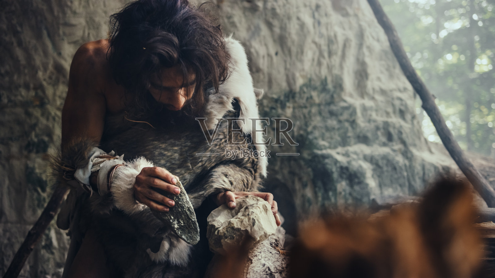 一个原始穴居人穿动物皮肤的特写镜头用锋利的石头击中岩石,使第一个原始工具狩猎动物猎物。尼安德特人使用燧石。人类文明的黎明。照片摄影图片