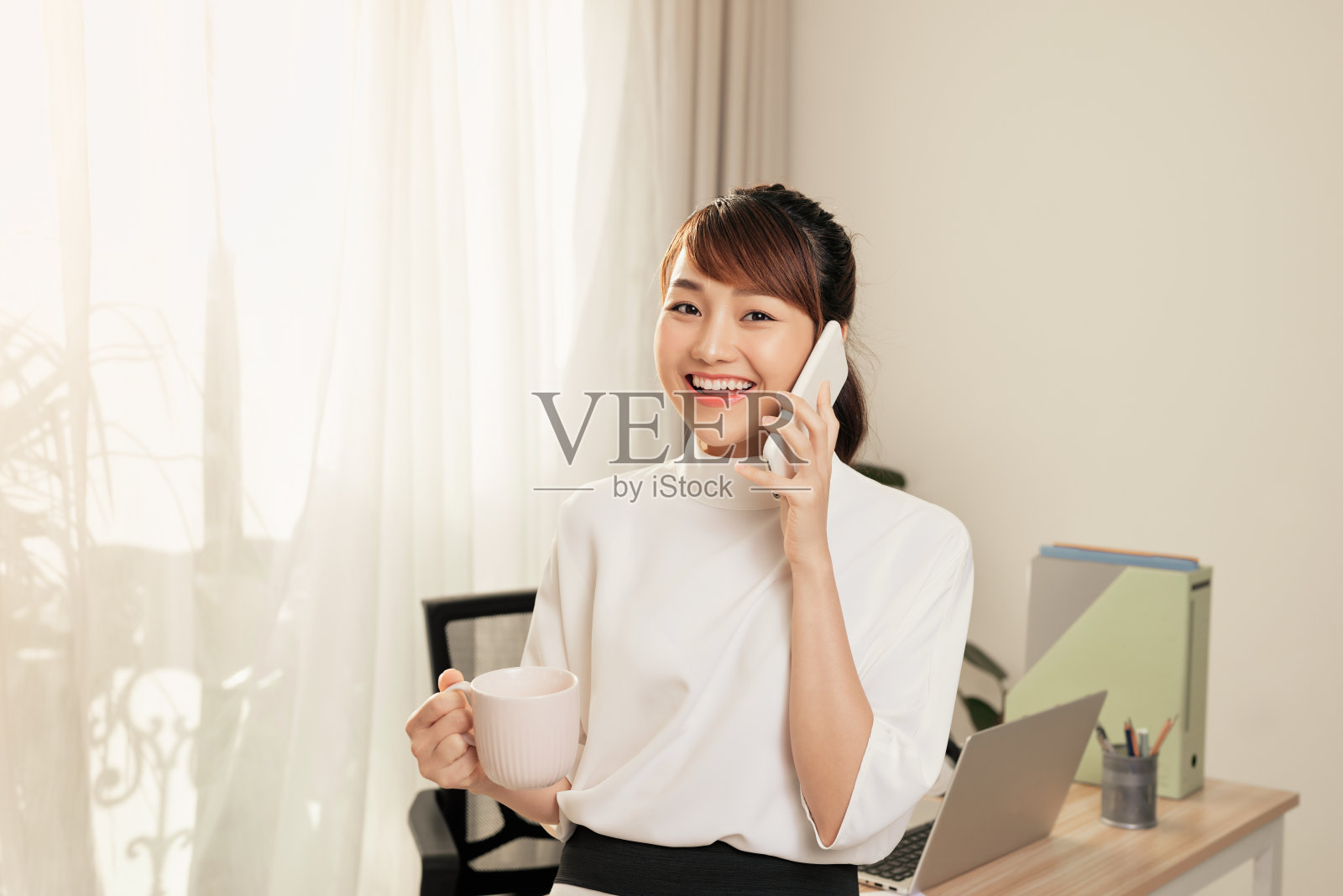 在办公室的桌旁喝咖啡时打电话的年轻亚洲女商人照片摄影图片