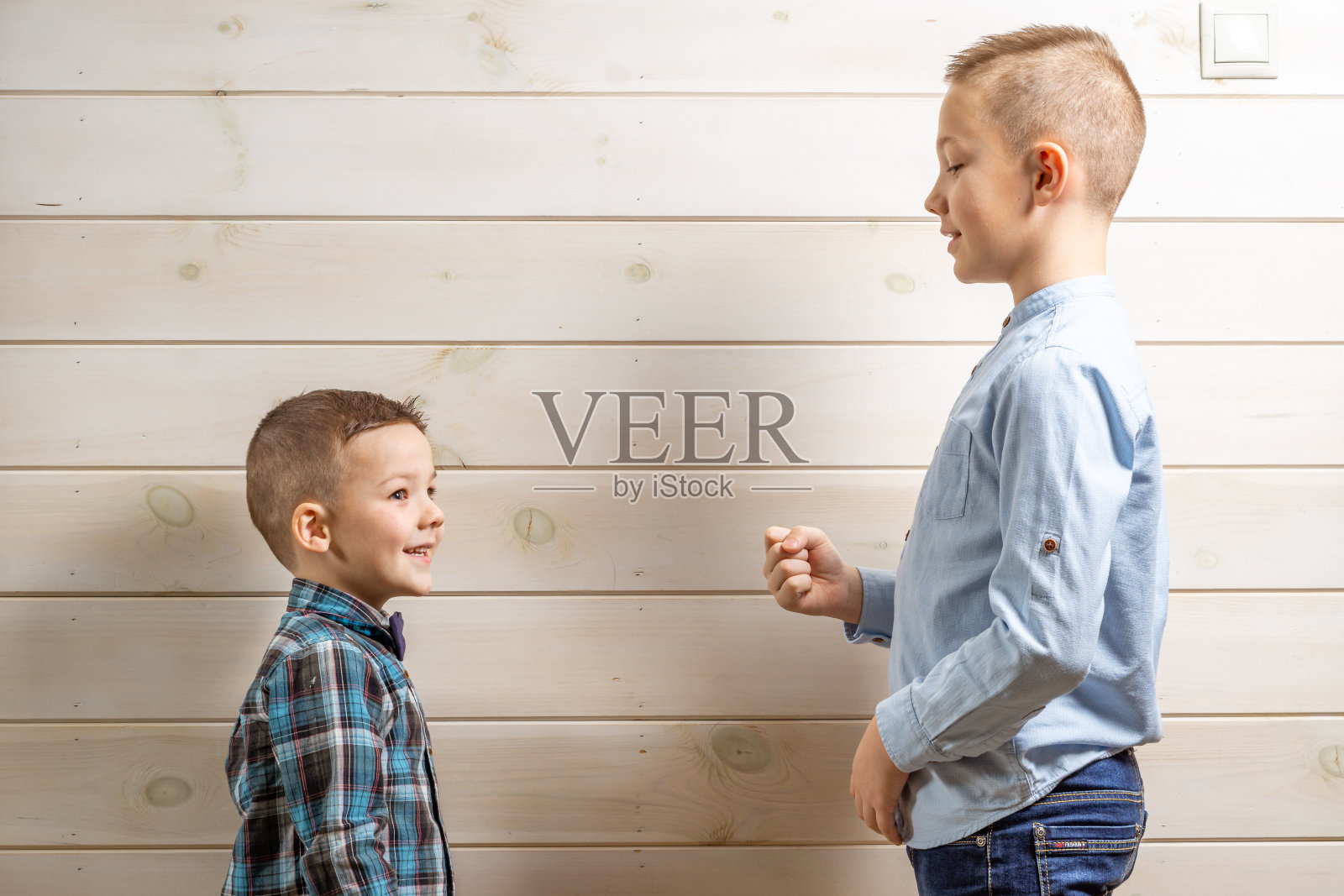 一个4岁的男孩穿着一件蓝色的偷窃衬衫,在一个浅色的木制背景下哭泣,他10岁的弟弟站在那里玩着石头剪刀布。照片摄影图片