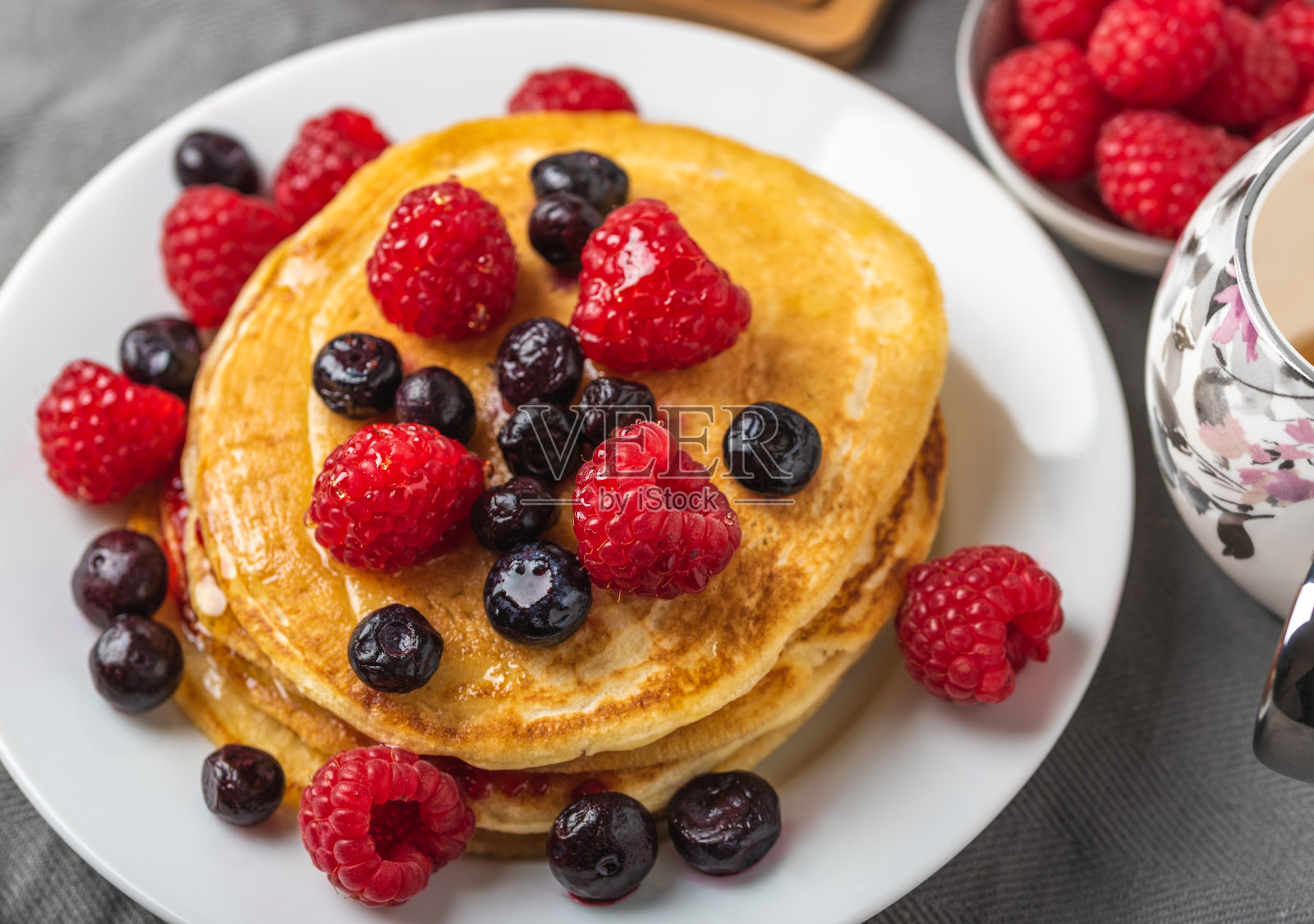 灰色的桌子上,白色的盘子里放着甜甜的自制煎饼和覆盆子。照片摄影图片