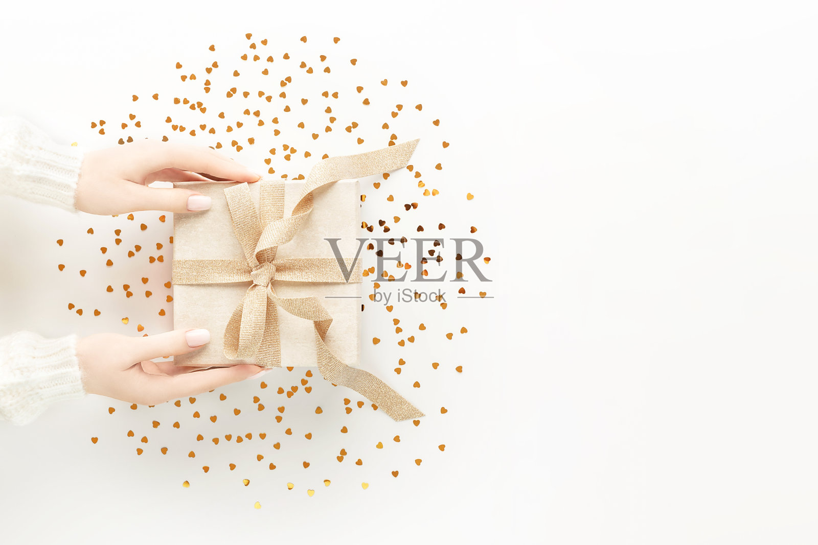 女性的手在温暖的白色毛衣与美丽的白色美甲持有优雅的金色蝴蝶结在白色背景与黄金心形闪闪发光的礼物盒。节日背景假日。平的风格。照片摄影图片