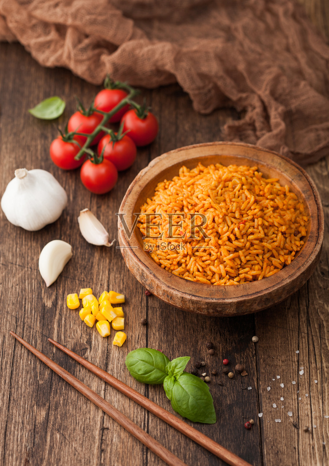 木碗里有煮熟的红色长谷物印度香米和蔬菜,木桌背景上有木棒和番茄、玉米、大蒜和罗勒。俯视图照片摄影图片