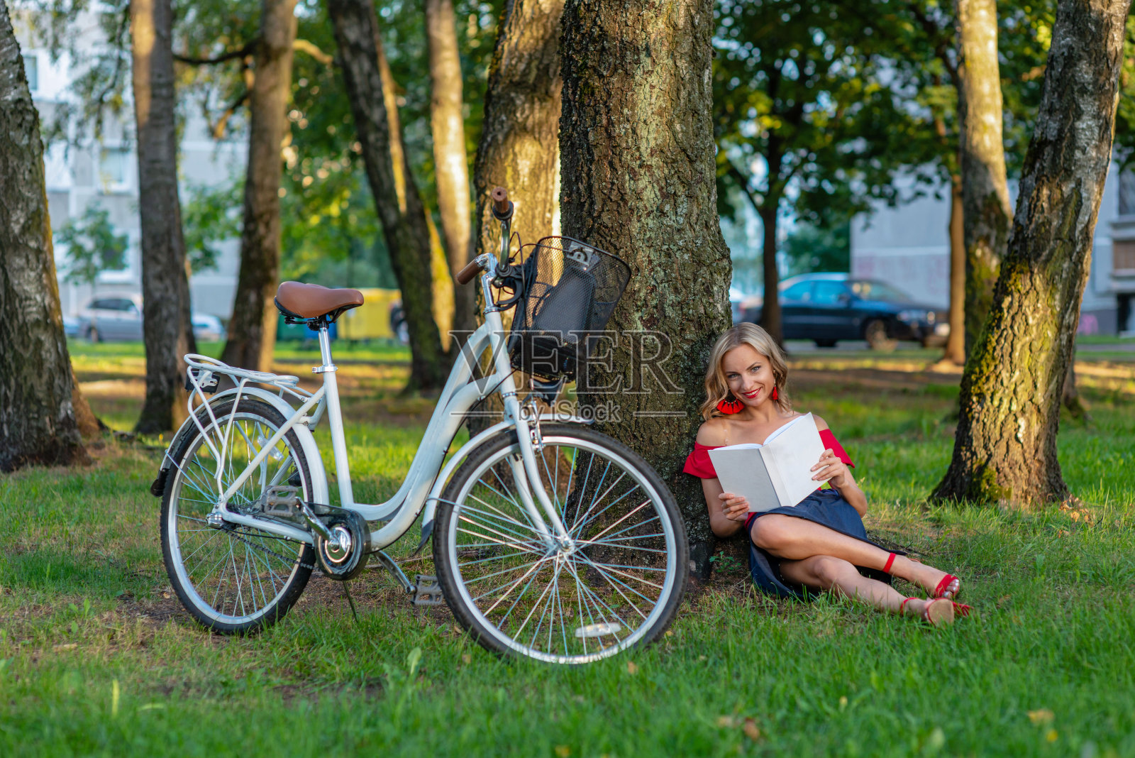 一个年轻的金发女人骑着一辆白色的自行车坐在公园里的一棵树旁,读着一本书——图片照片摄影图片