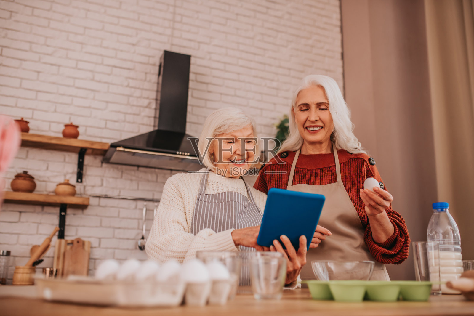 头发花白、面带微笑的女士们穿着围裙,读着新的食谱照片摄影图片