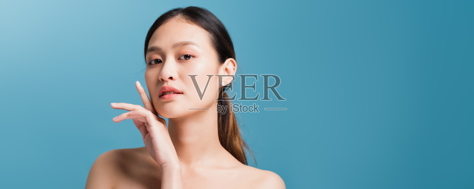 自信美丽的亚洲女性,手放在脸旁,看着相机,在蓝色背景下拍摄的肖像。健康面部肌肤护理概念。照片摄影图片