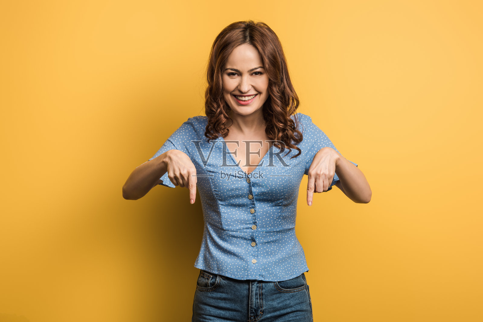 在黄色背景上,快乐的女人看着镜头,用手指指向下方照片摄影图片