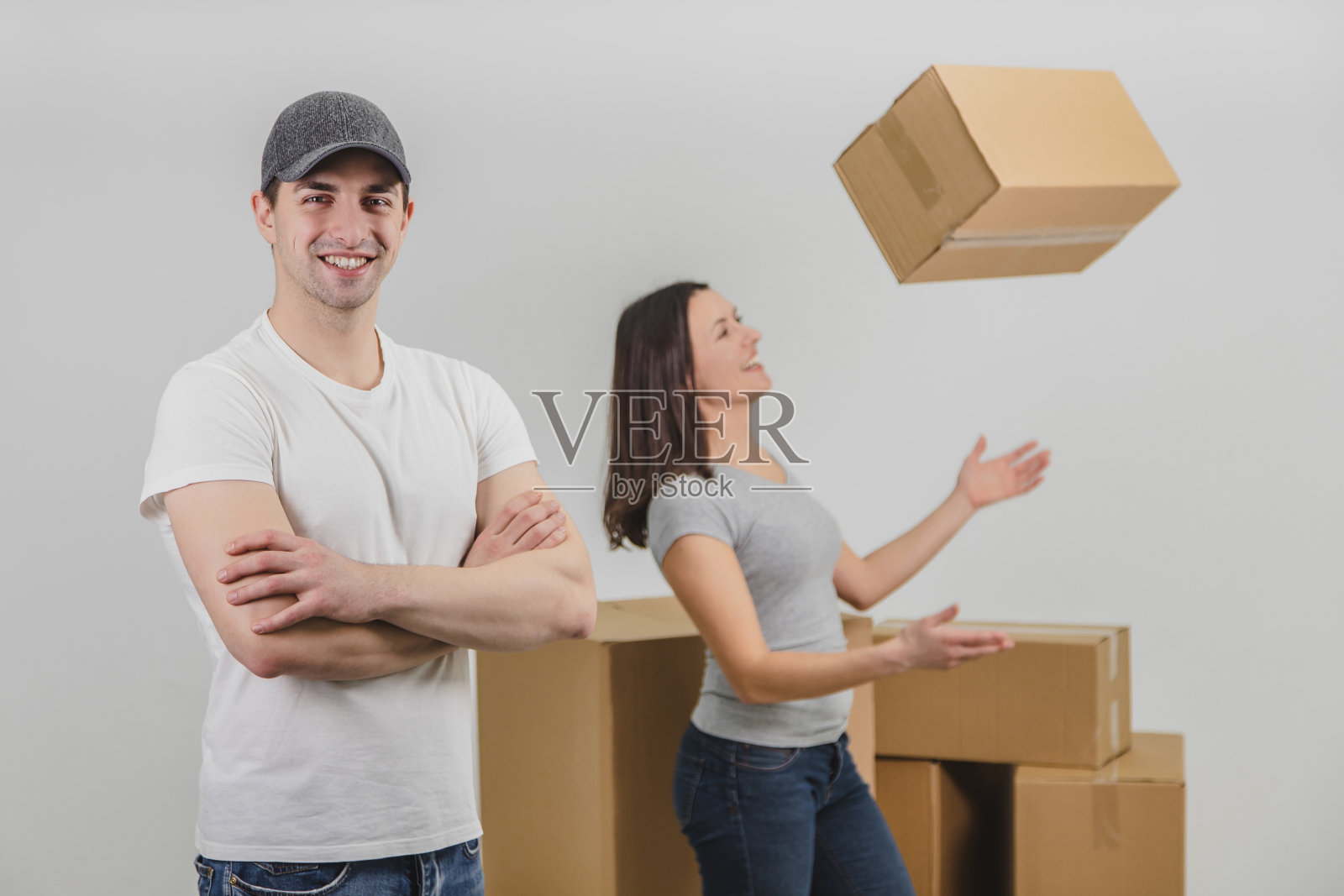 强壮的快递员把箱子运到了可爱的年轻女子的新公寓,他站在最前线,双手交叉。一个高兴的女人正站在背景上,模糊地抛起盒子。照片摄影图片