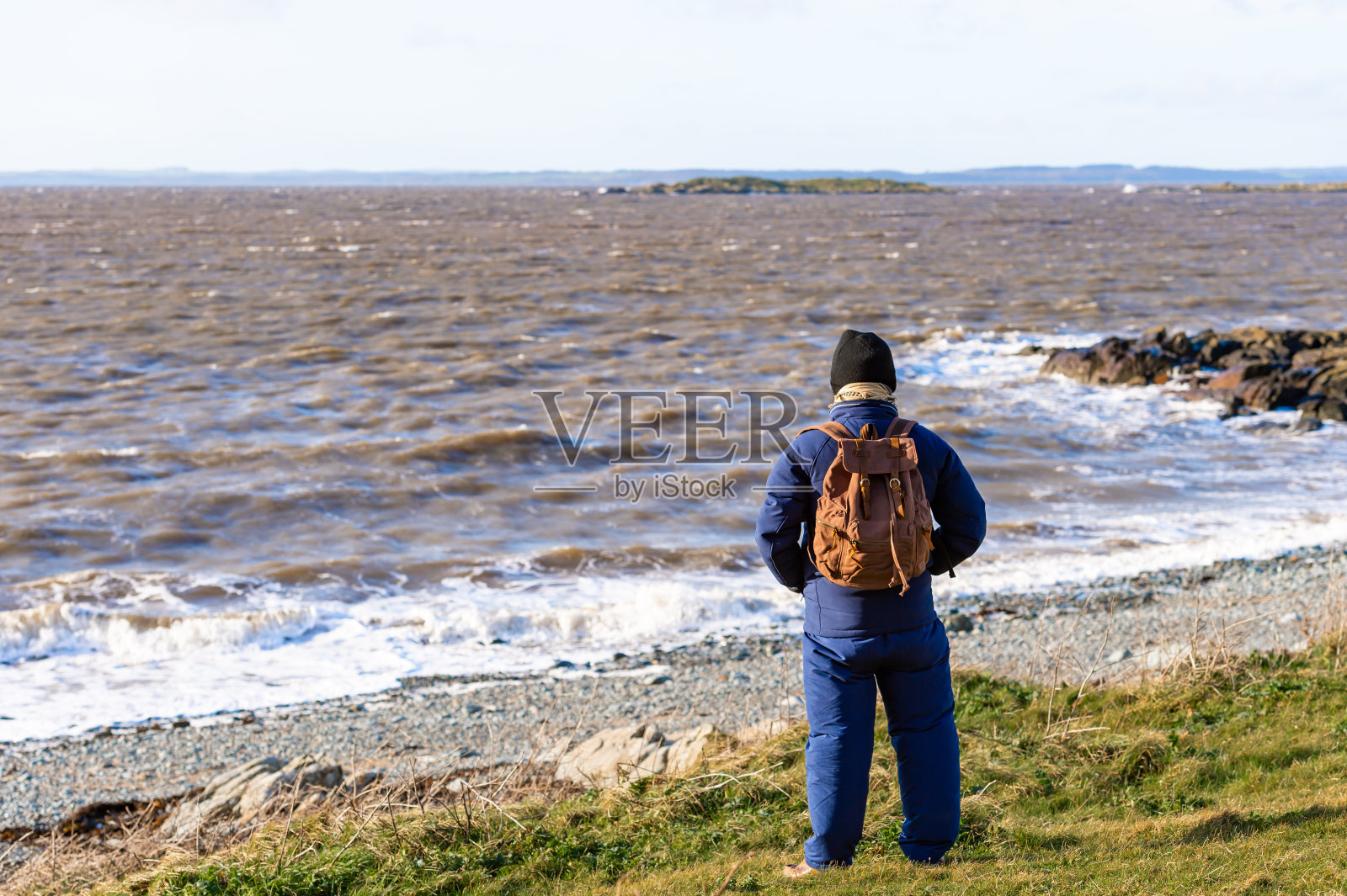 一个老人站在多岩石的海岸上,这是一个晴朗的冬天的早晨,涨潮了。照片摄影图片