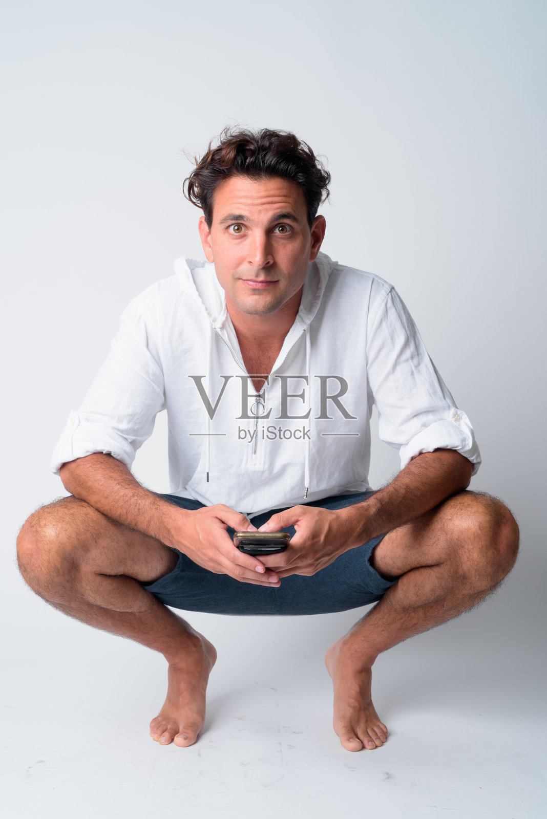 全身拍摄的英俊的西班牙男子蹲下,而持有电话照片摄影图片