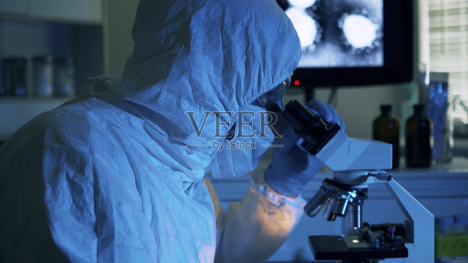 手持电脑显示器上显示冠状病毒图像的场景,身着防护服的科研人员使用显微镜分析血液样本。照片摄影图片