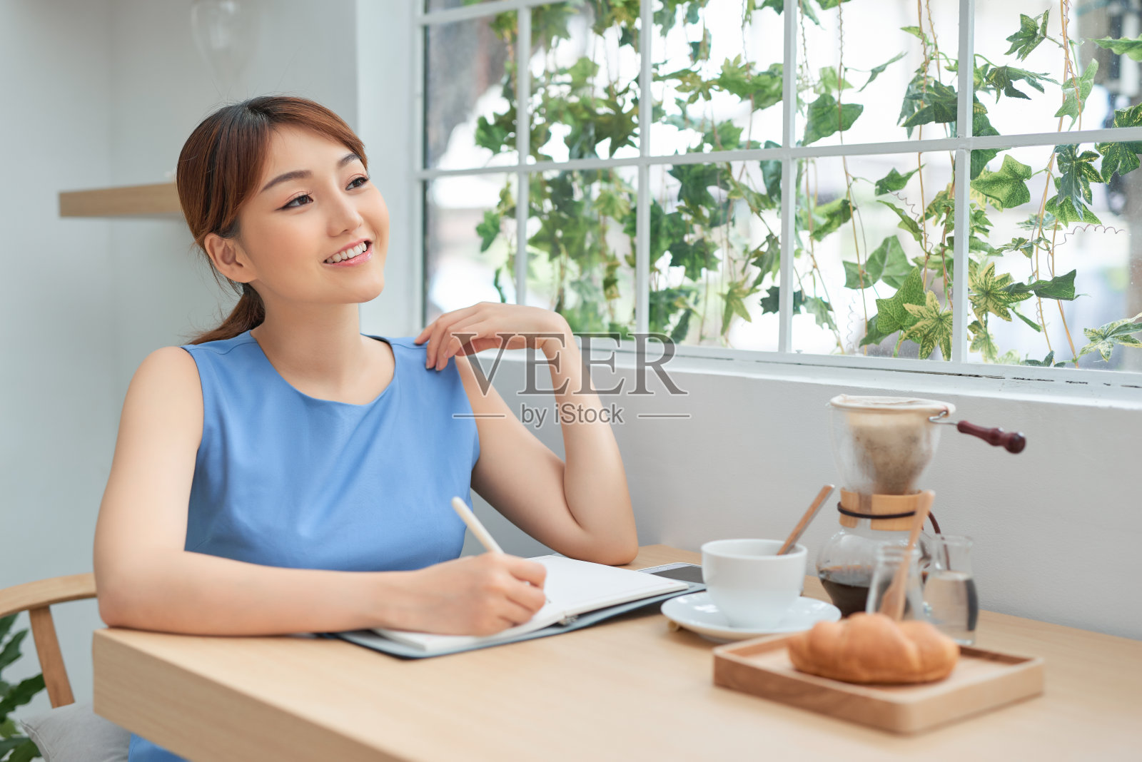 坐在窗后,一边用手机一边在笔记本上书写的年轻亚洲女性照片摄影图片