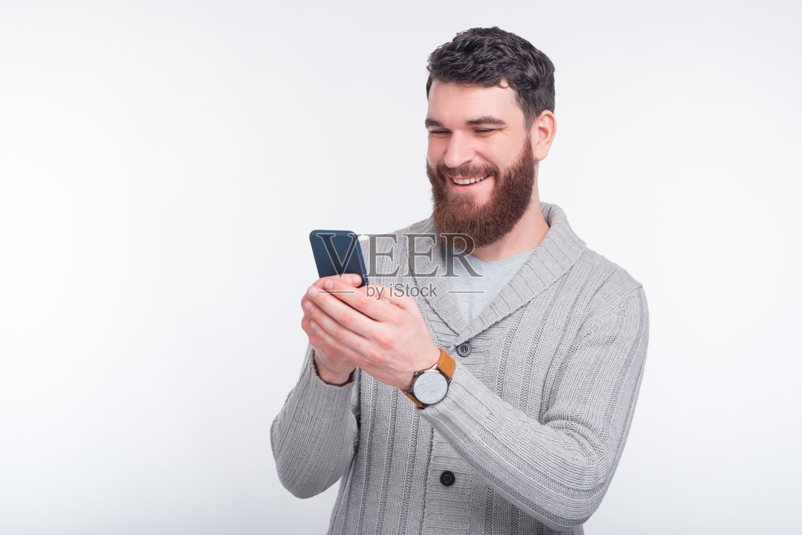 快乐的大胡子男人正在他的深蓝色手机上输入一些东西在白色的背景。照片摄影图片