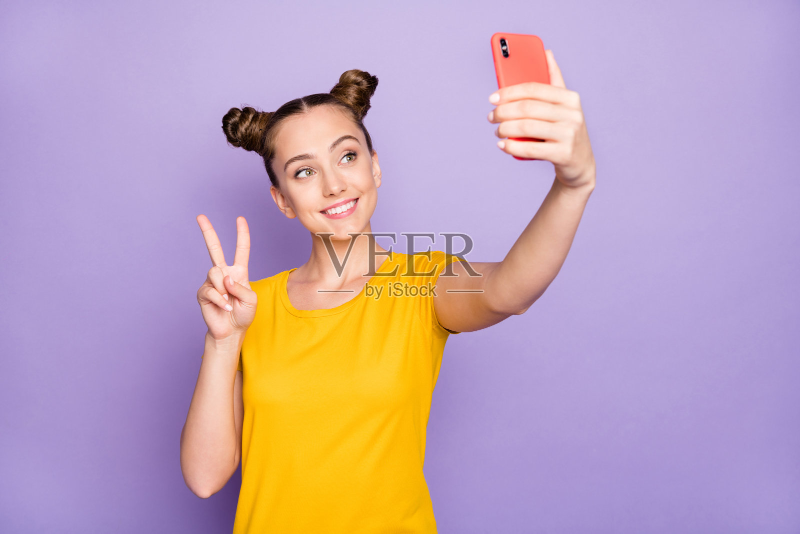 漂亮女士拿着电话,展示V形手势,为社交博客自拍,穿着黄色T恤,淡紫色背景照片摄影图片
