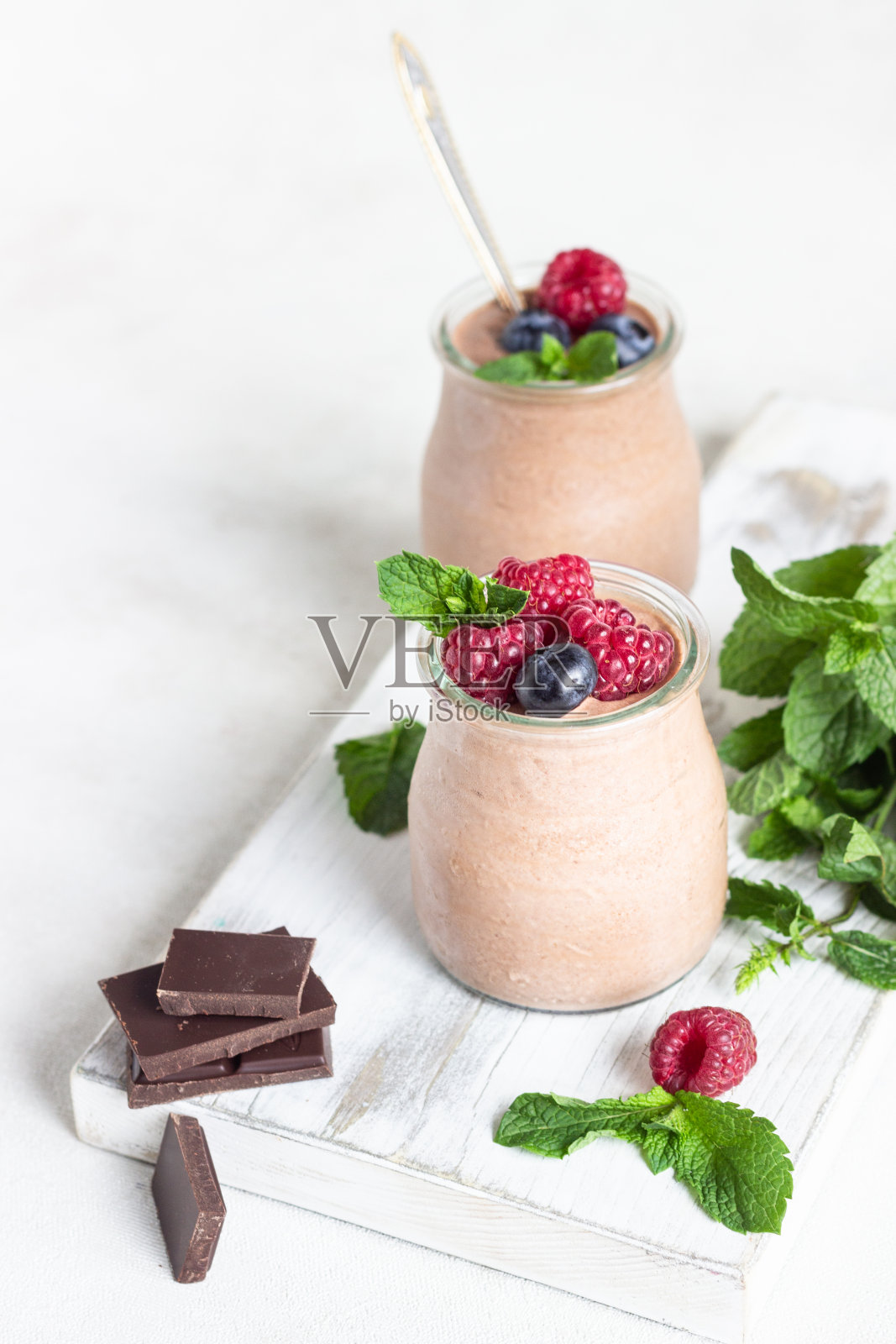 巧克力甜点(意式奶冻,慕斯或布丁),装在玻璃罐中,配上覆盆子,蓝莓和薄荷,背景为浅灰色。照片摄影图片