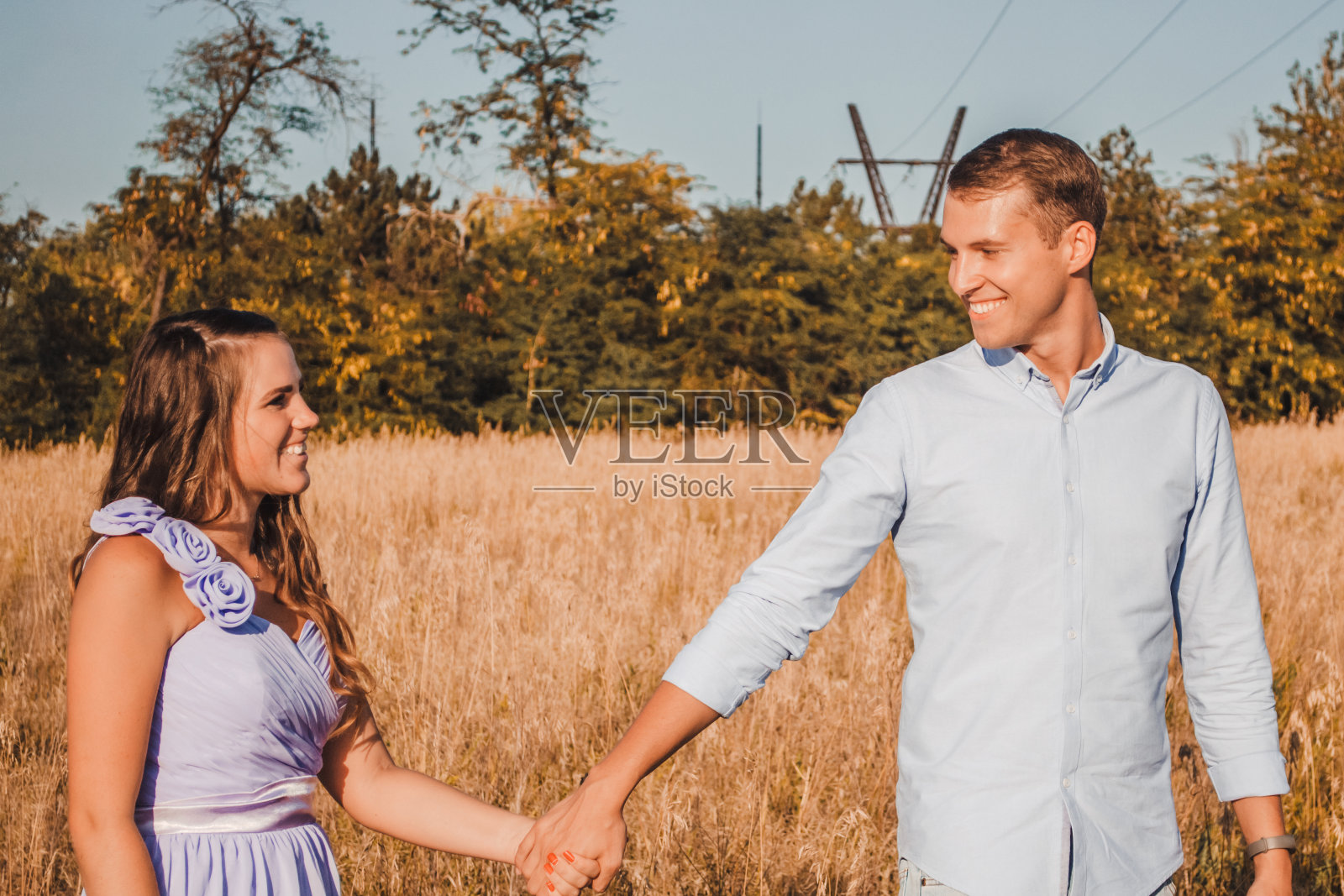 年轻情侣手牵手在干草地里微笑。男人带领女人前进。穿着浅色衣服的美丽浪漫情侣在夏天摆姿势的户外肖像照片摄影图片