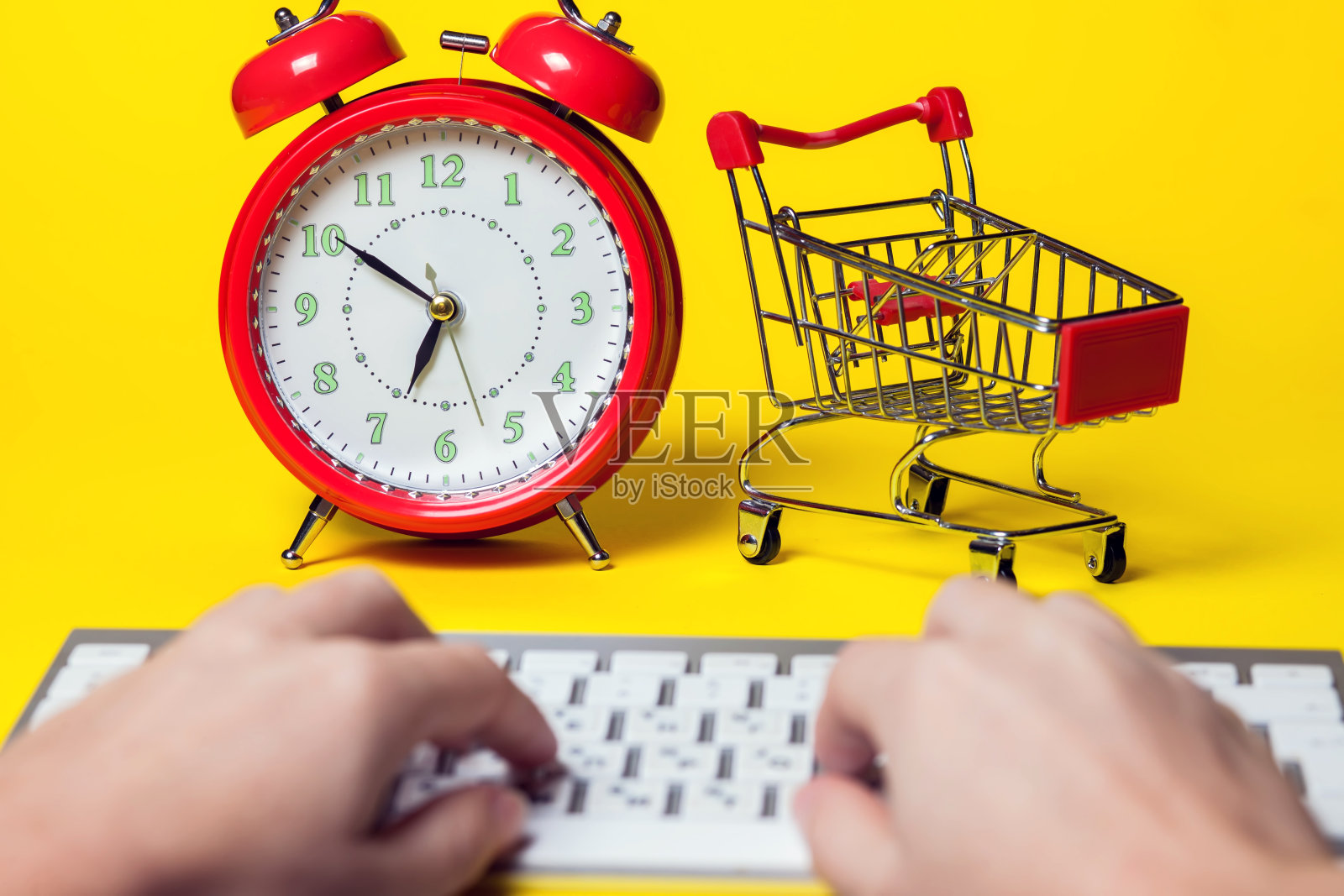 闹钟、小型购物车、在彩色背景上使用键盘的手。购物概念照片摄影图片