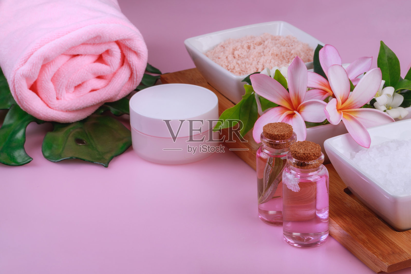 水疗用品组合:碗里装着白色和粉色岩盐和鲜花,粉色背景上的毛巾、乳霜和乳液照片摄影图片