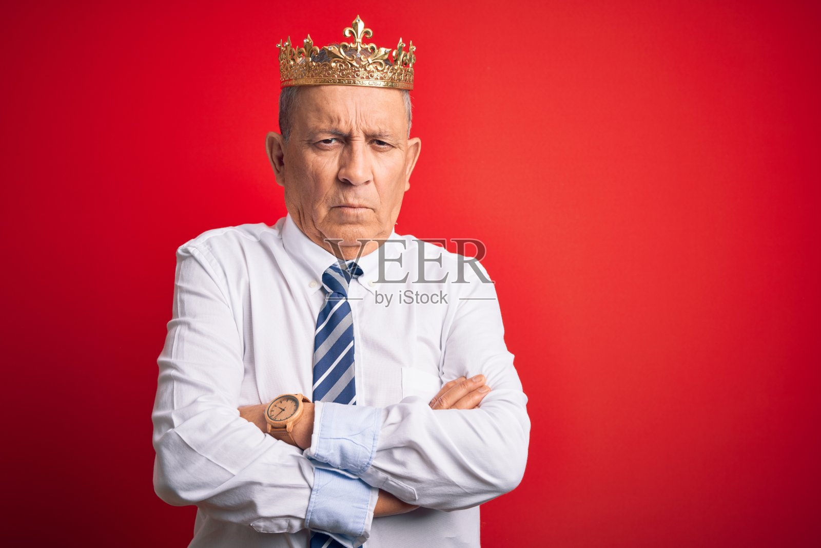 年长的英俊商人戴着国王皇冠站在红色背景前,持怀疑态度和紧张,脸上带着不赞成的表情,双臂交叉,是个消极的人照片摄影图片