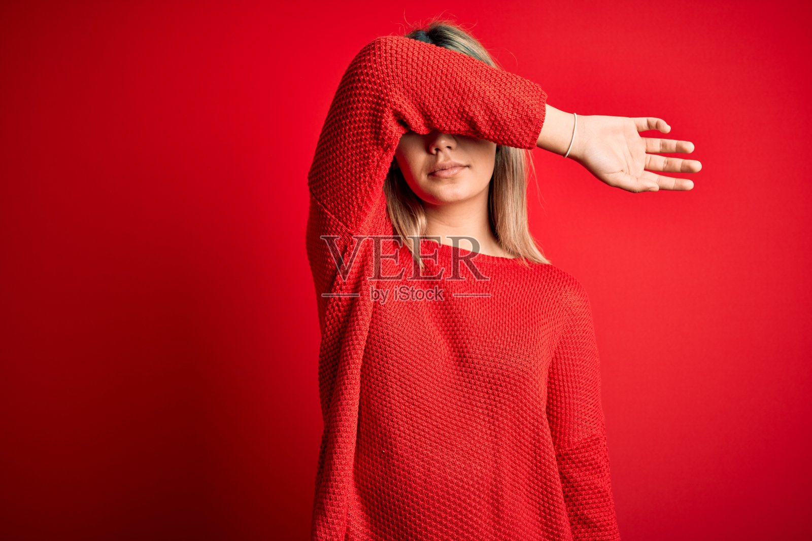 年轻金发女人穿休闲毛衣,用手臂遮眼,表情严肃悲伤。照片摄影图片