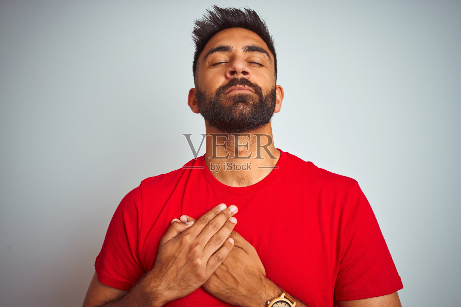 身穿红色T恤的年轻印度男子,在白色背景前闭着眼睛,双手放在胸前,面带感激的微笑照片摄影图片