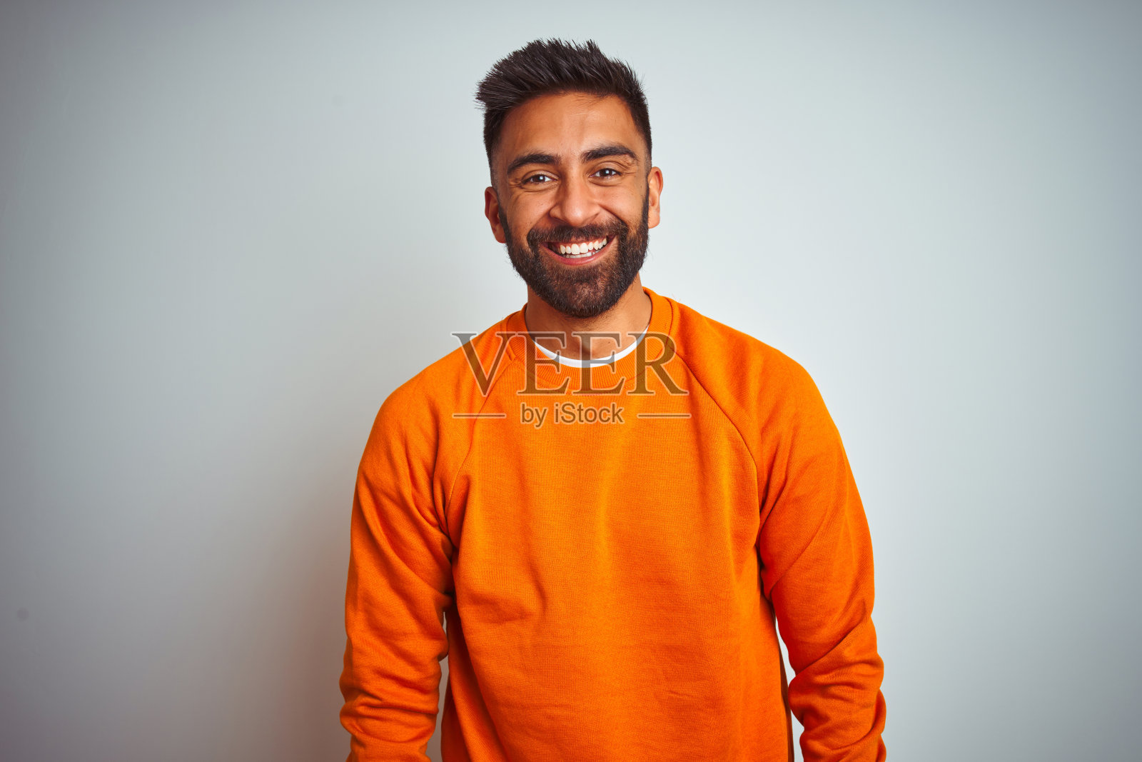 穿着橙色毛衣的年轻印度男子,在白色背景前露出幸福而酷的笑容。照片摄影图片