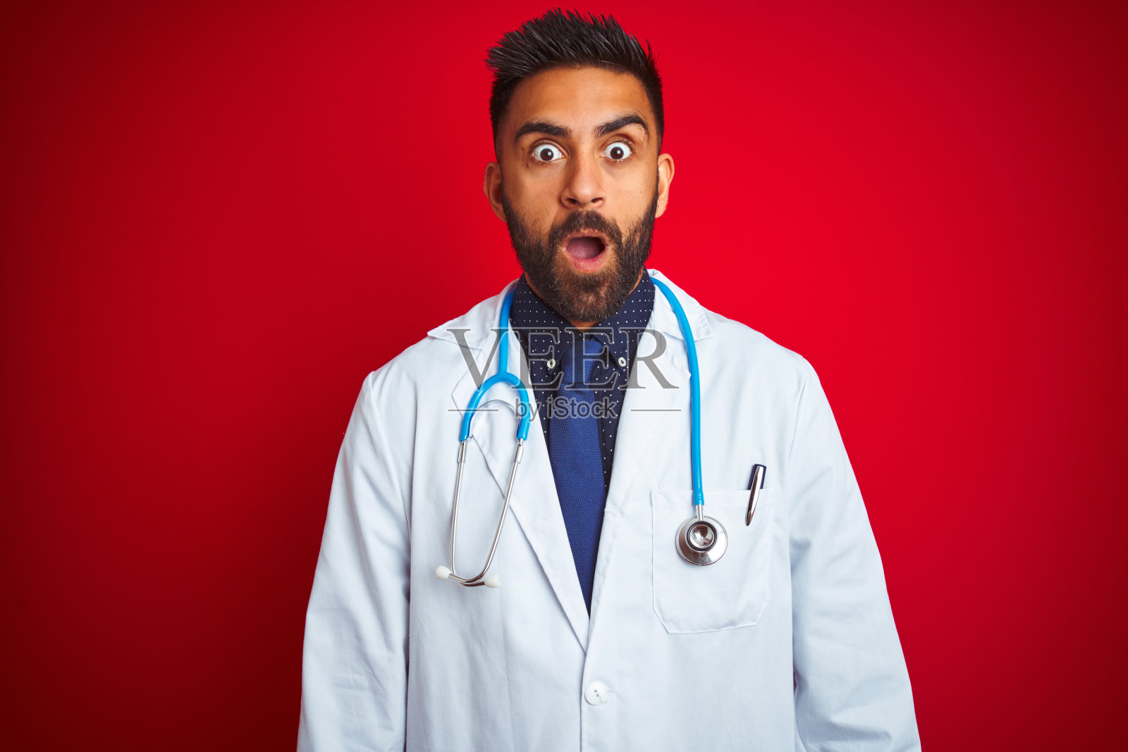 年轻的印度男医生戴着听诊器站在红色背景前,面露惊讶、恐惧和兴奋的表情。照片摄影图片