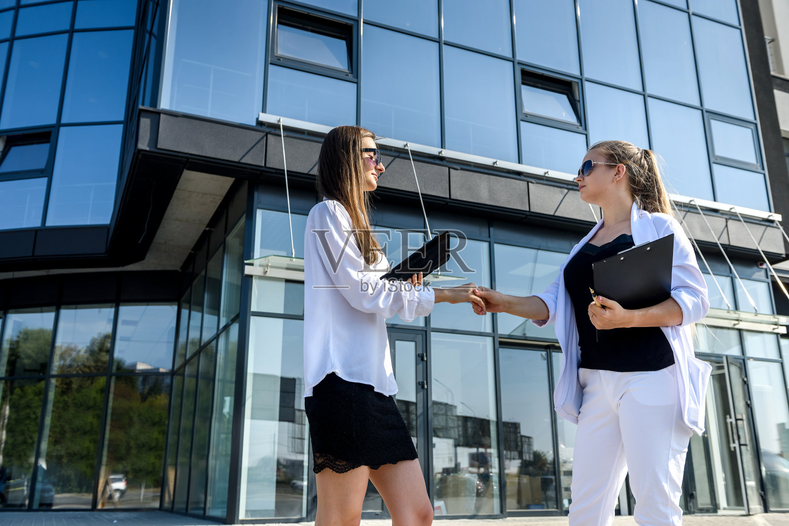 商务合作伙伴在办公楼前握手,两位穿着商务套装的美丽女性互相微笑照片摄影图片