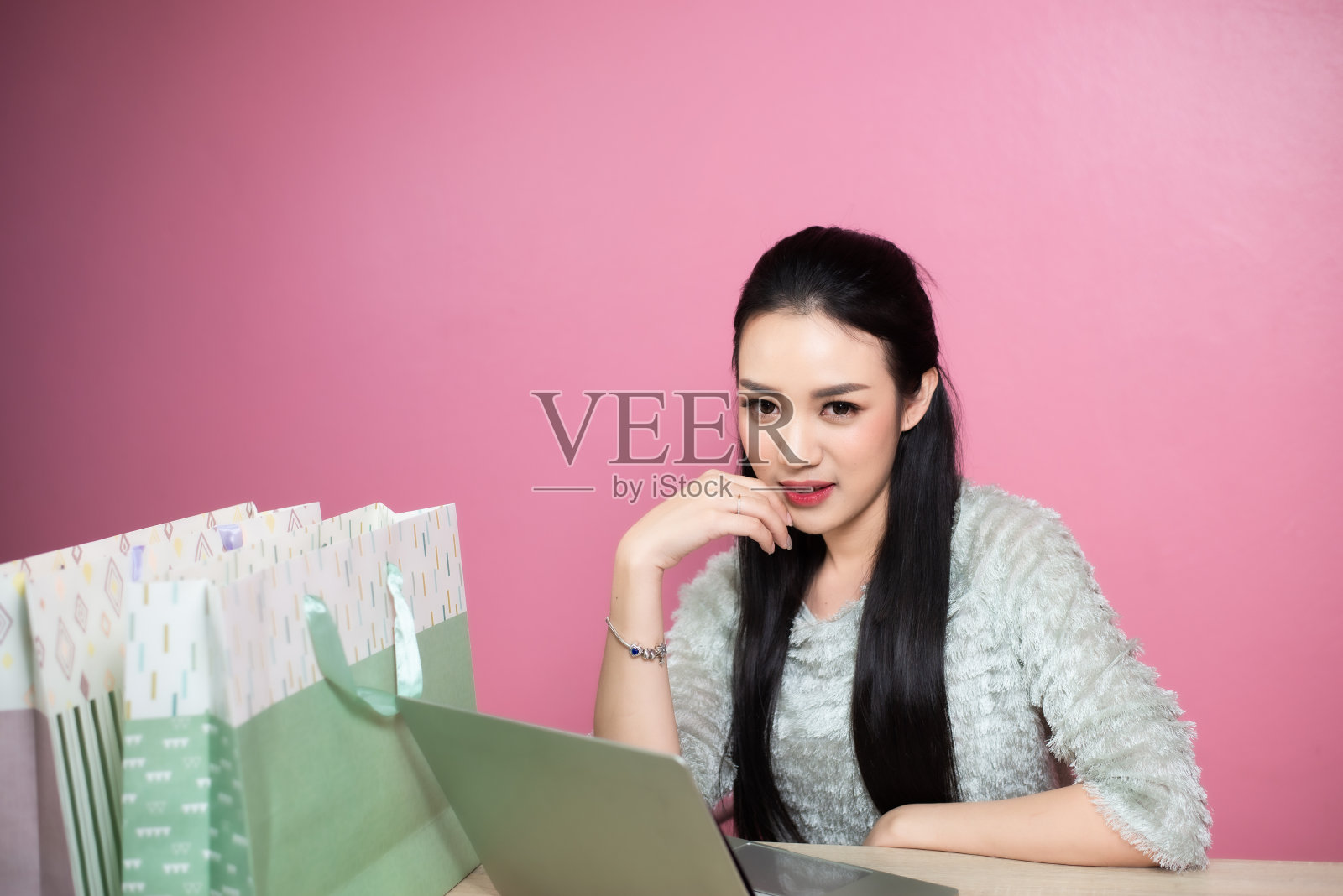 美丽亚洲微笑的女人用笔记本电脑购物袋在桌子上粉红色的背景。照片摄影图片