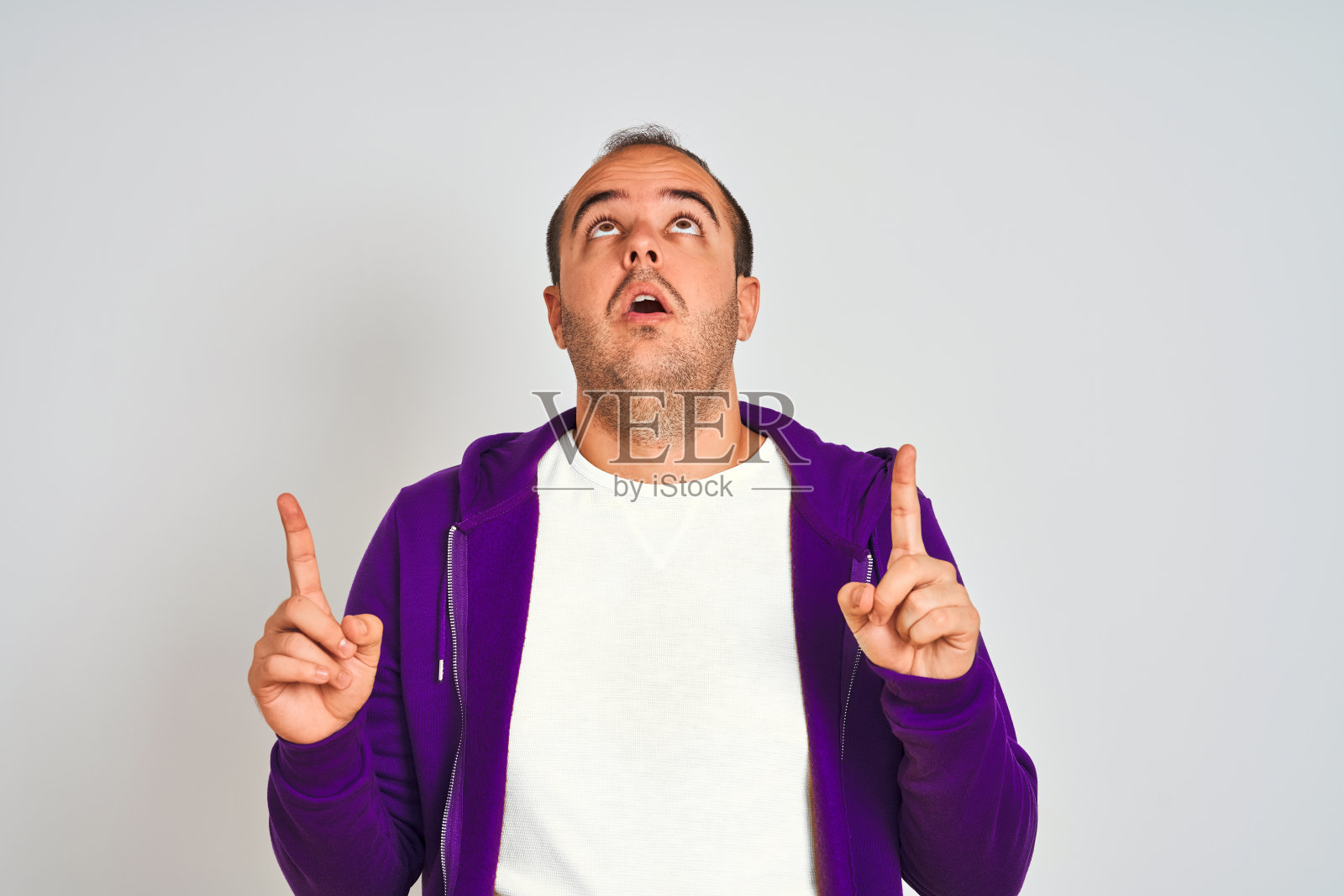 一名身穿紫色运动衫的年轻人站在孤立的白色背景下,惊讶地抬起头,用手指指着前方。照片摄影图片