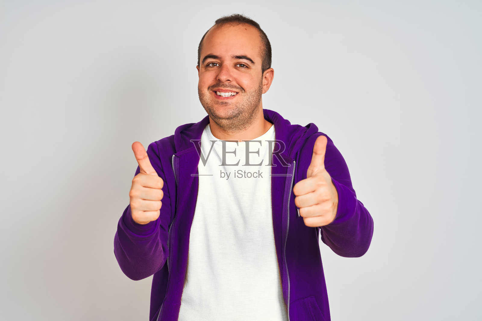 年轻男子穿着紫色的运动衫站在孤立的白色背景下,用手做积极的手势,竖起大拇指微笑着,为成功而高兴。赢家的姿态。照片摄影图片