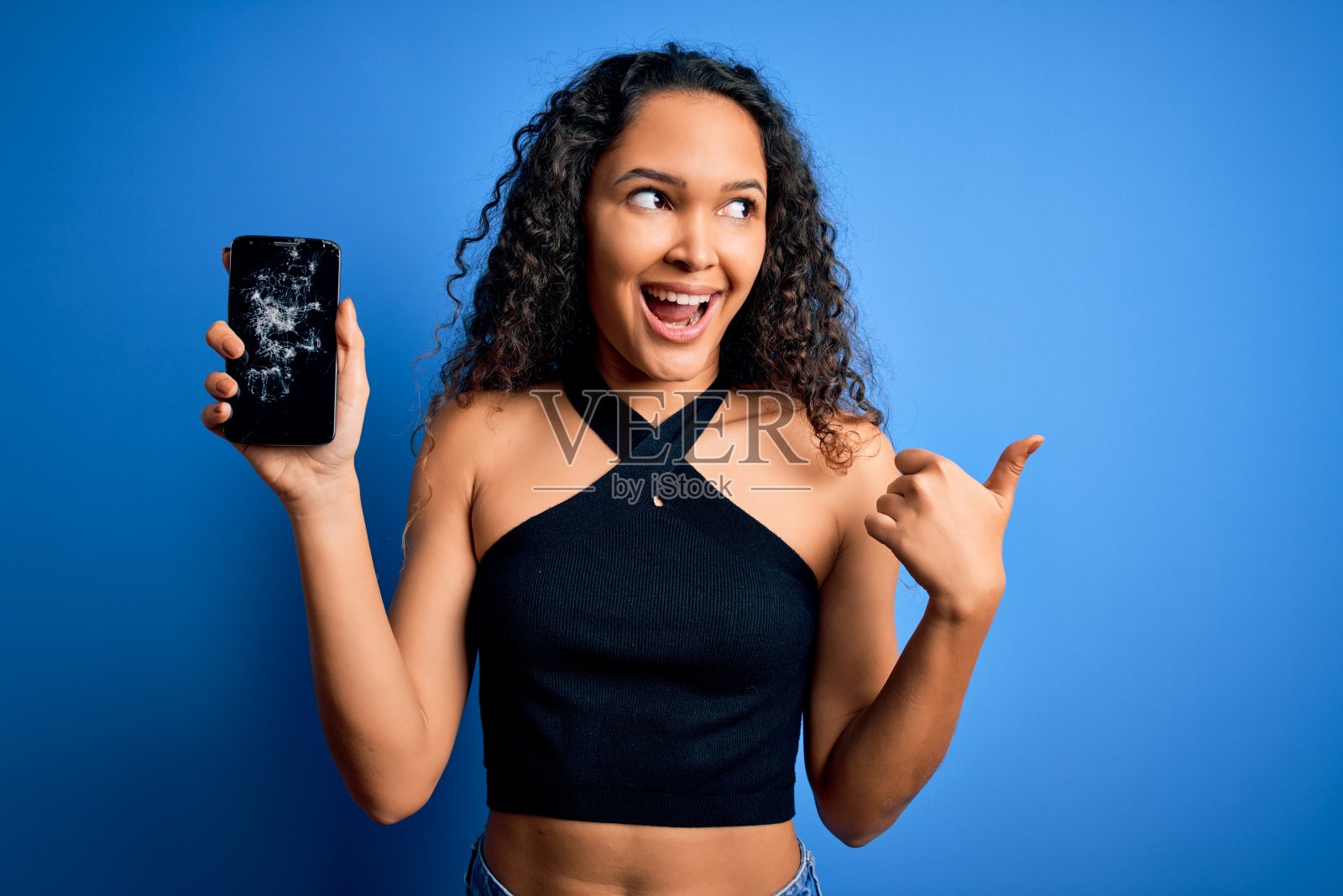 年轻美丽的女人卷发拿着破碎的智能手机显示破碎的屏幕指向和显示拇指向上的一边与幸福的脸微笑照片摄影图片