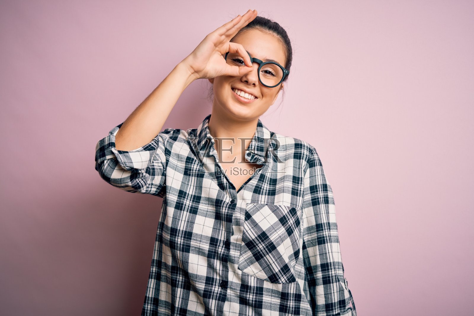 年轻美丽的黑发女人穿着休闲衬衫和眼镜在粉红色的背景做ok手势与手微笑,眼睛透过手指与快乐的脸。照片摄影图片