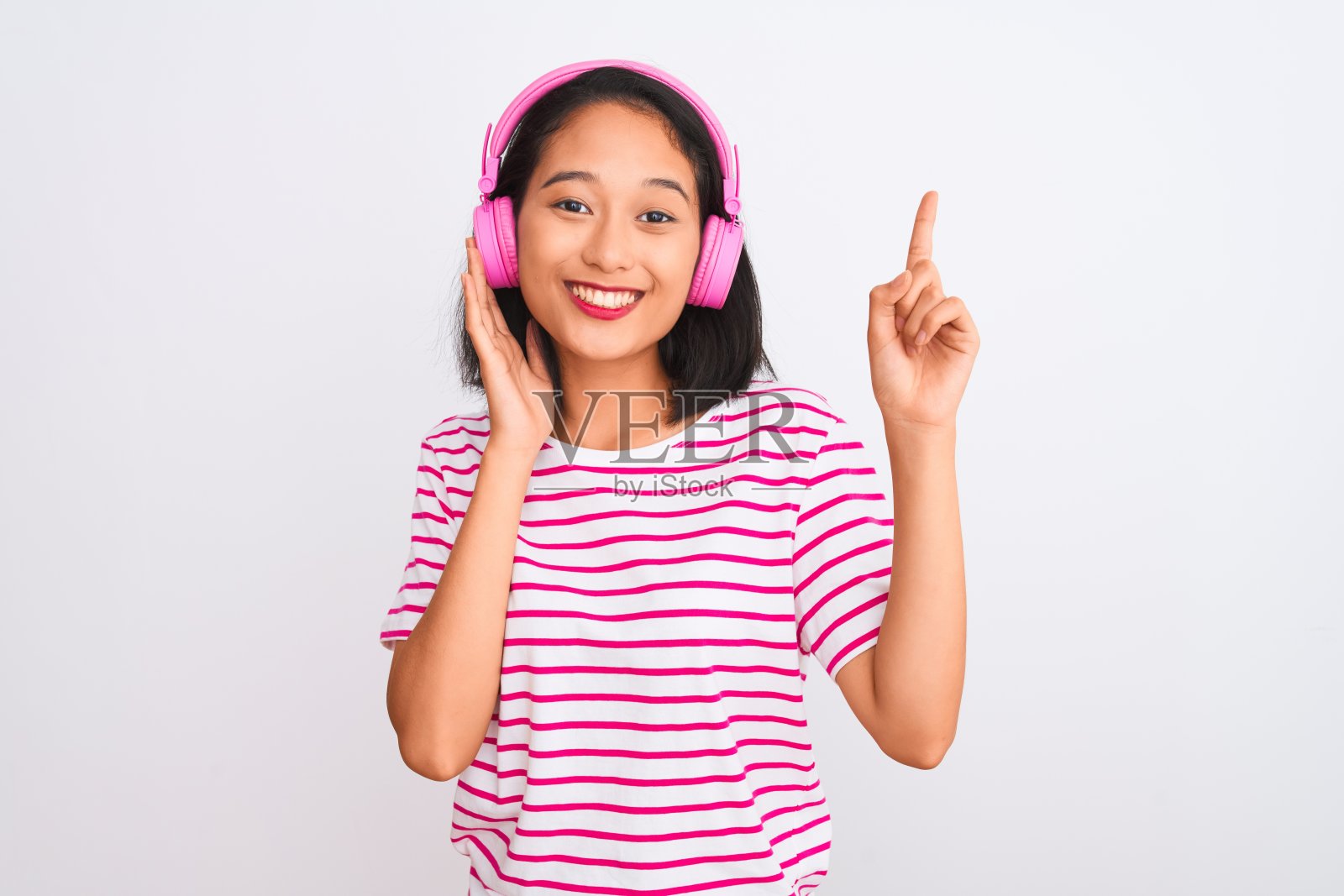 美丽的中国女人在孤立的白色背景下戴着耳机听音乐,惊讶地想到一个想法或问题,开心地指着对方的脸,第一照片摄影图片