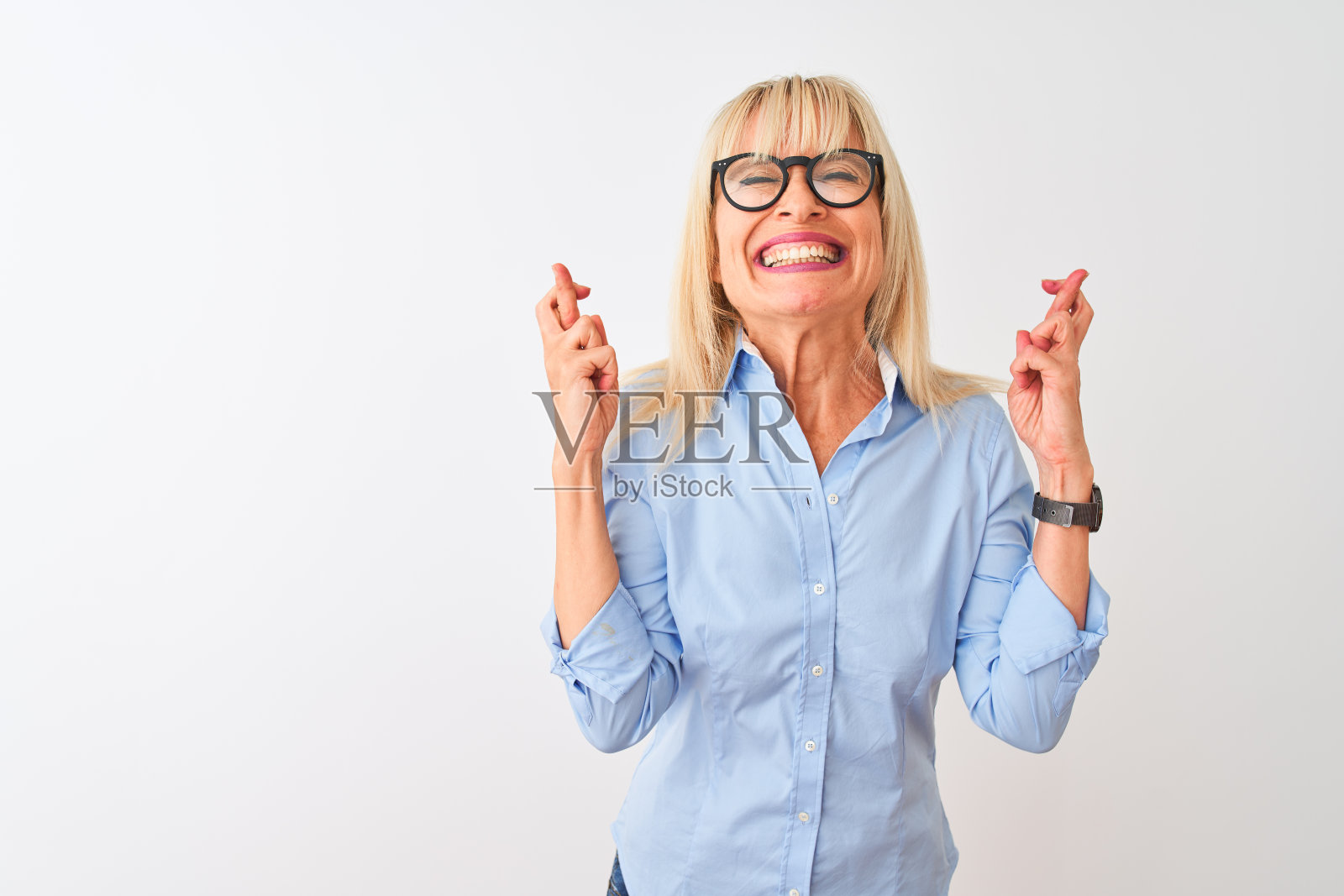 中年女商人穿着优雅的衬衫和眼镜,在白色背景前比划着手指交叉,面带微笑,闭着眼睛,表达希望。幸运和迷信的概念照片摄影图片