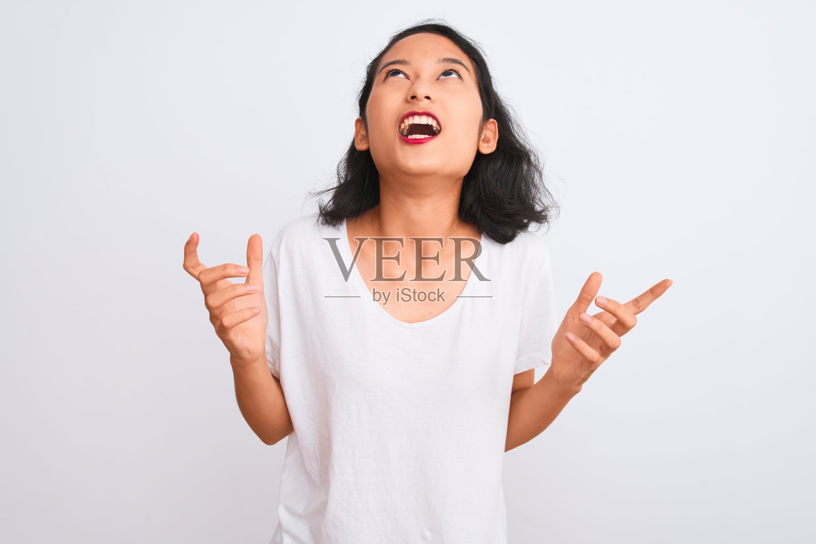 年轻的中国女人穿着休闲t恤站在孤立的白色背景疯狂地大喊大叫,带有攻击性的表情和举起手臂。挫折的概念。照片摄影图片