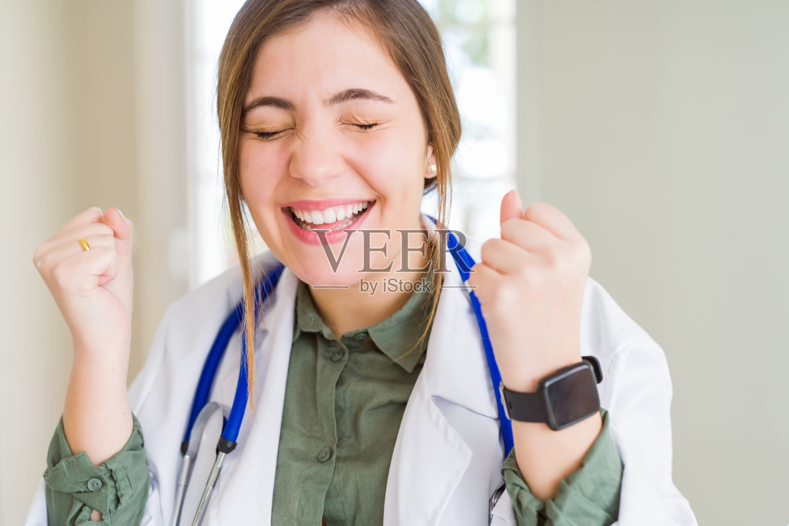 年轻漂亮的女医生身穿白大褂,戴着听诊器,举起双臂庆祝胜利,笑容满面,充满成功的喜悦照片摄影图片