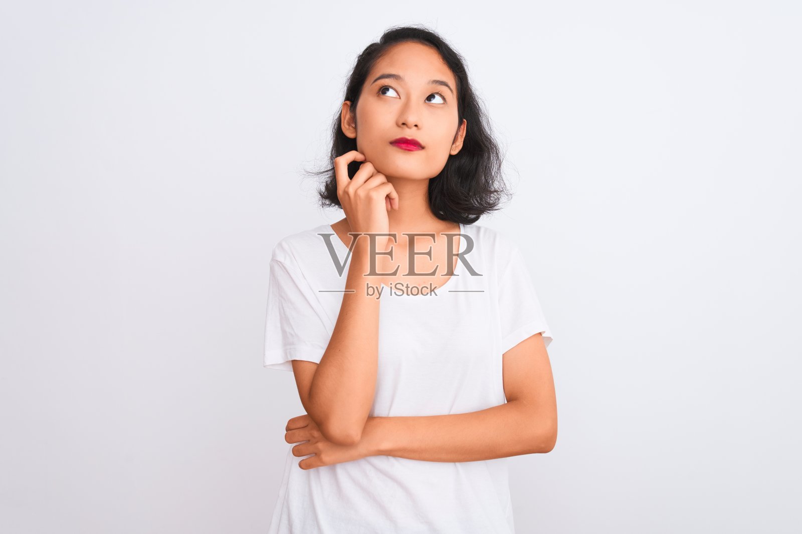 年轻的中国女子穿着休闲t恤站在孤立的白色背景下,手托着下巴思考问题,沉思的表情。带着沉思的微笑。怀疑的概念。照片摄影图片