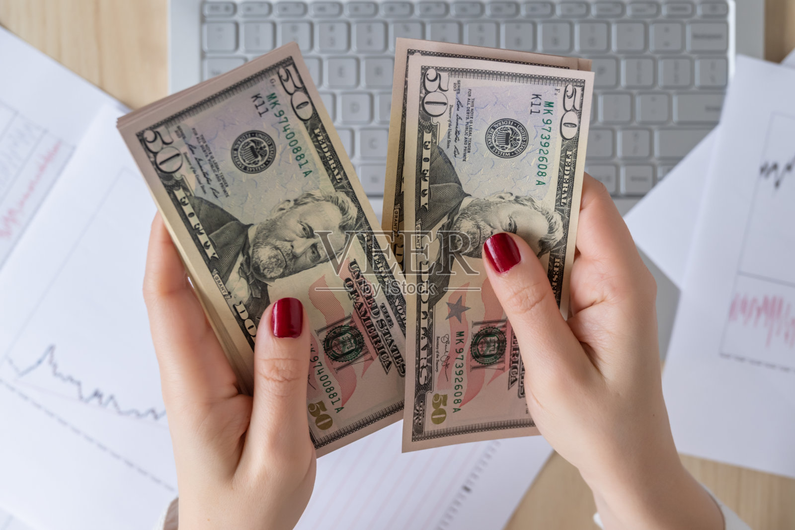 时间流逝。女性自由职业者手里拿着50美元钞票,计算着利润和收入。将图形和图表打印在办公桌上的纸上的笔记本电脑。平躺照片摄影图片