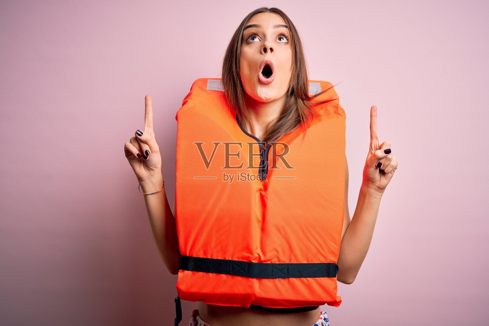 年轻美丽的褐发女人穿着橙色的救生衣在孤立的粉红色背景惊讶和惊讶的看着和指着手指和举起的手臂。照片摄影图片