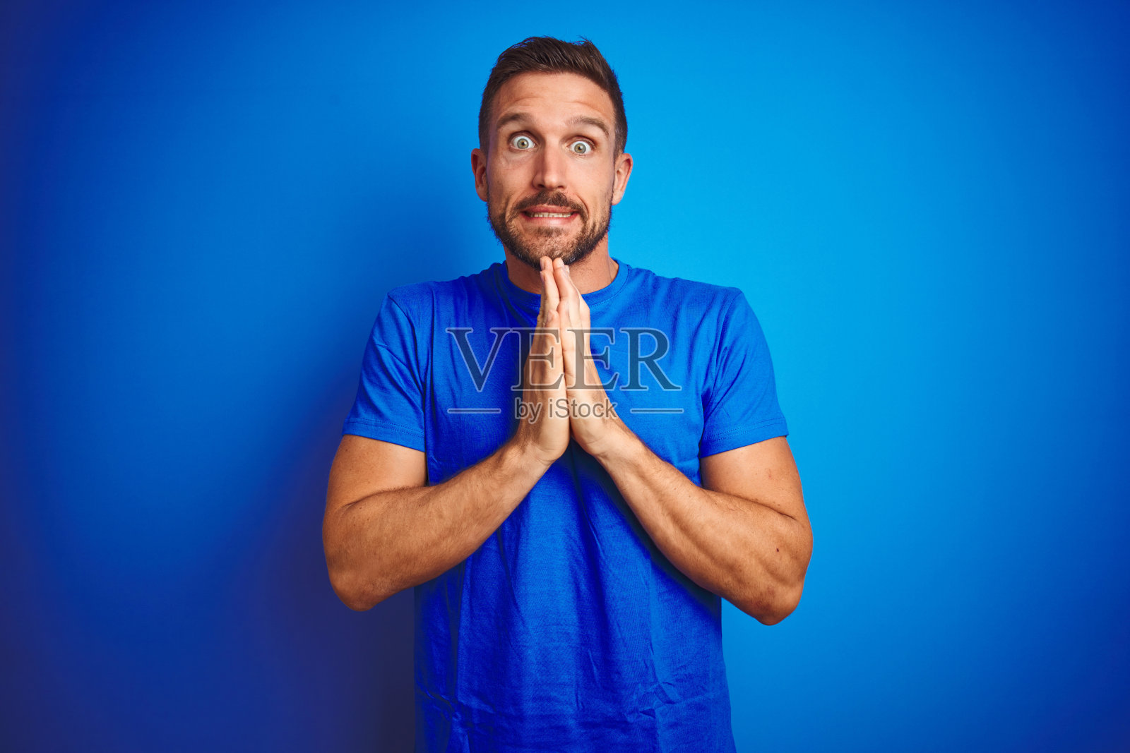 年轻帅气的男人穿着休闲T恤,在蓝色背景下双手合十祈祷,面带自信的微笑照片摄影图片