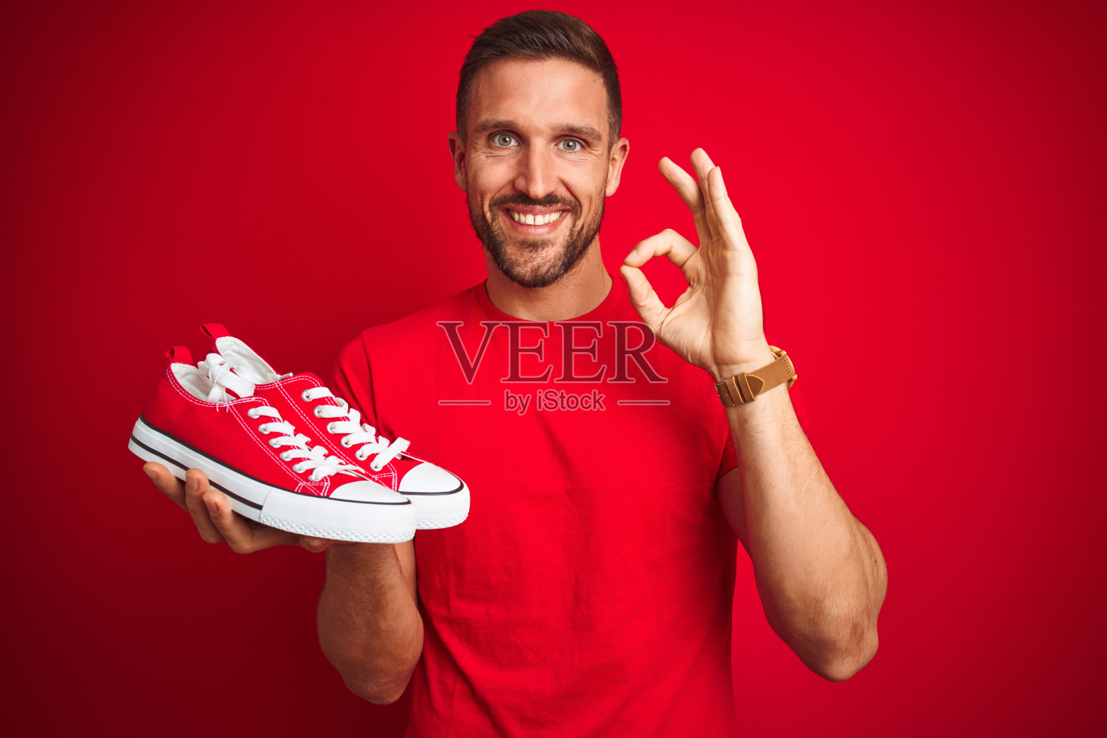年轻男人拿着休闲运动鞋,在红色背景前做出OK手势照片摄影图片