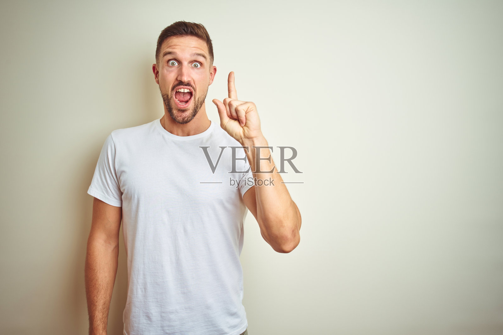 穿着休闲白色T恤的年轻英俊男人在背景前竖起手指,表示有了成功的想法,兴奋和快乐,第一照片摄影图片